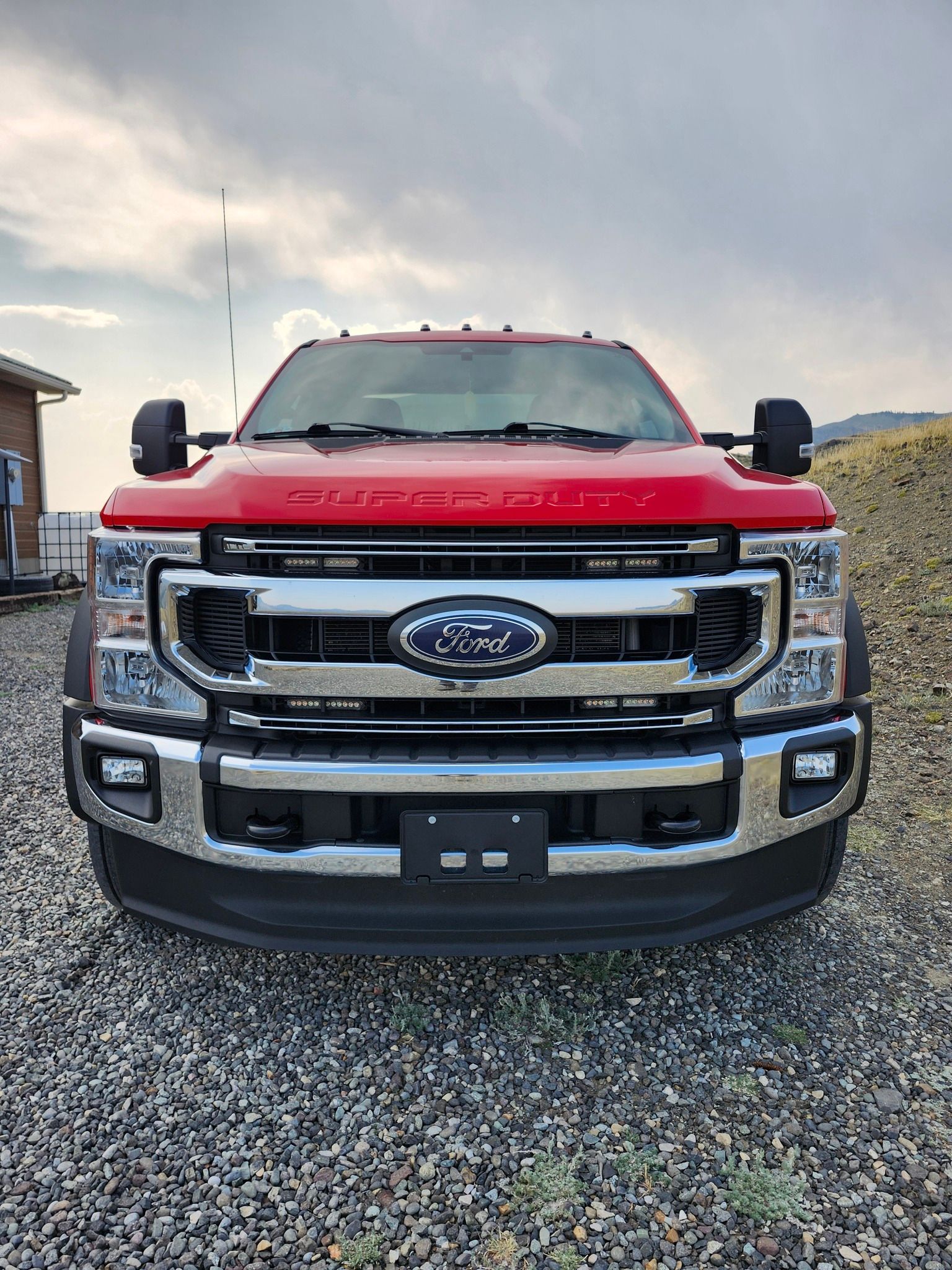 Red Ford Super Duty truck parked outdoors in front of gravel and a building, with a cloudy sky overhead.