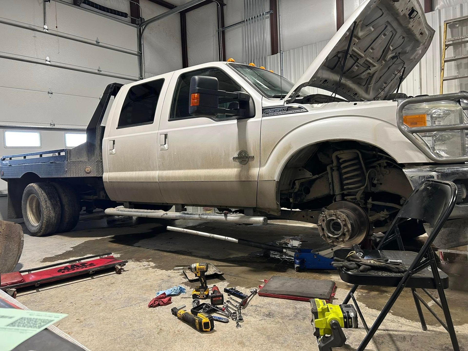 A white work truck being repaired in a garage, with the hood open and front wheel removed.