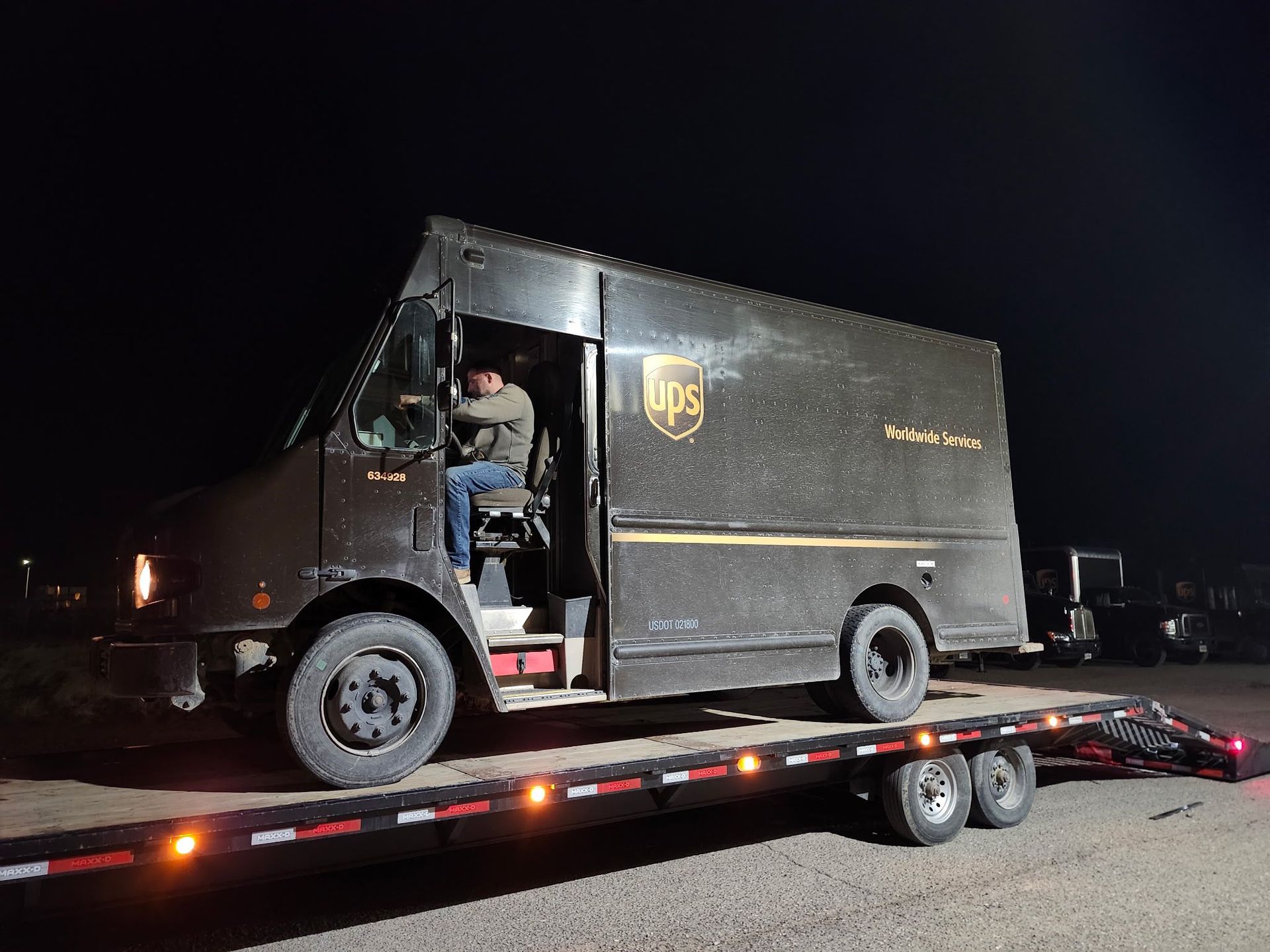 UPS truck on a trailer at night with a driver visible inside. The truck is brown with the UPS logo.