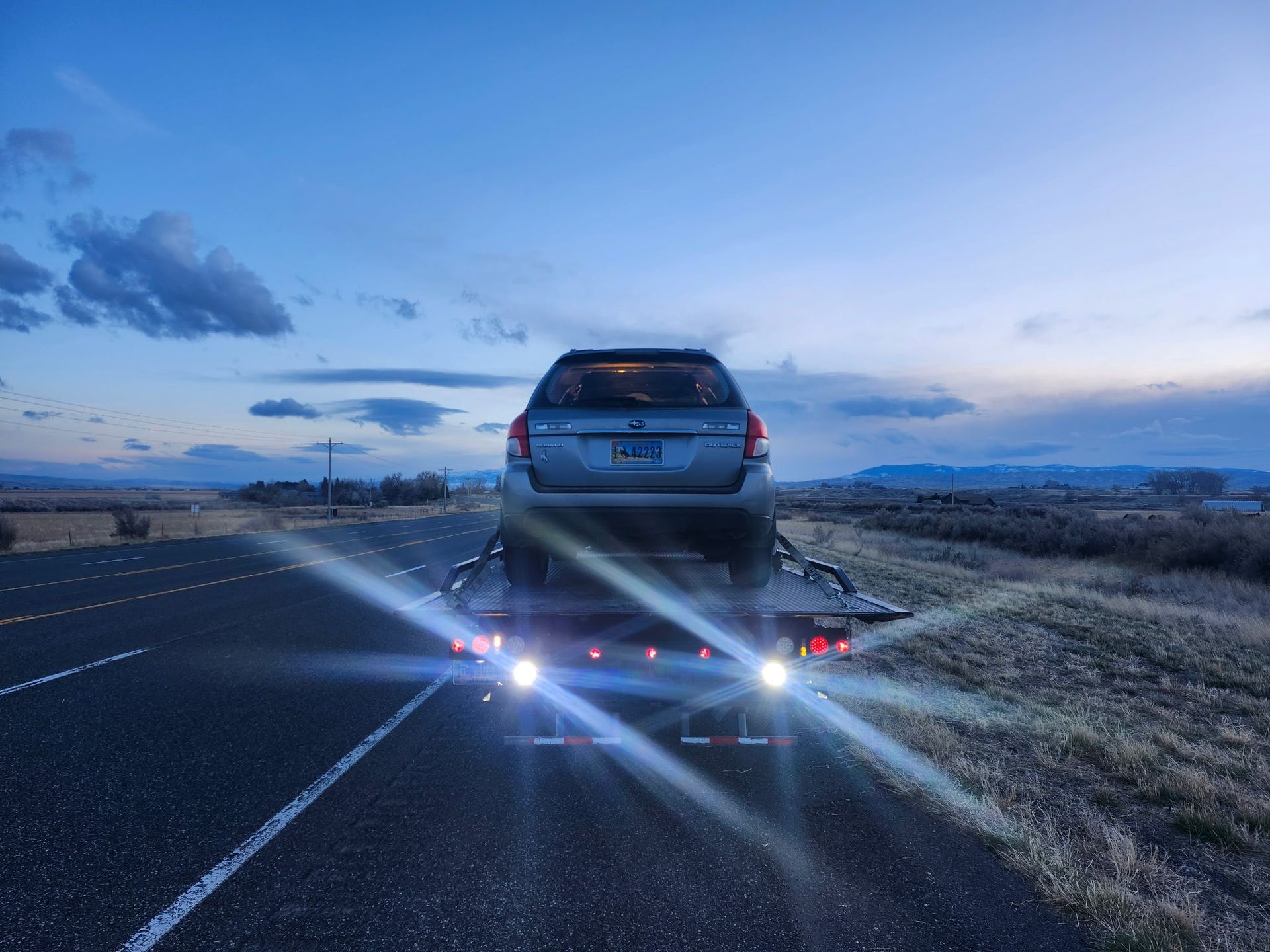 A silver car is on a trailer, seen from behind on a road. The setting is dusk or dawn, with fields and a cloudy sky.