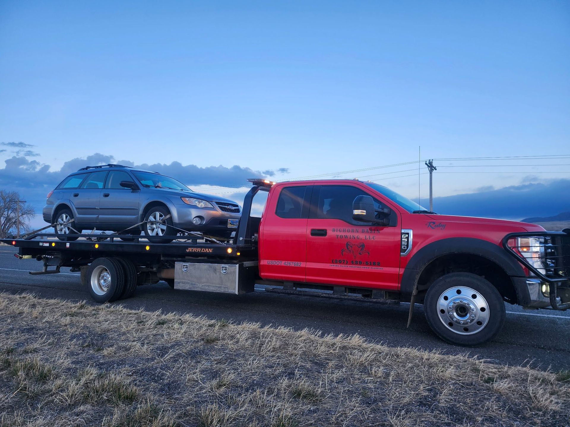 Red tow truck carrying a gray car on a flatbed. Sunset in the background.