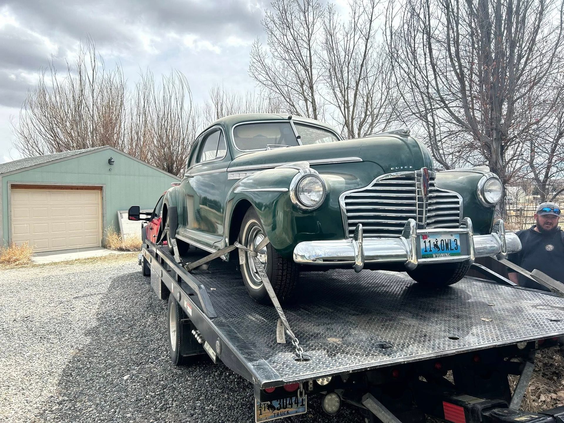 A vintage green Buick car being towed on a flatbed truck in front of a building with a garage door, outdoors.