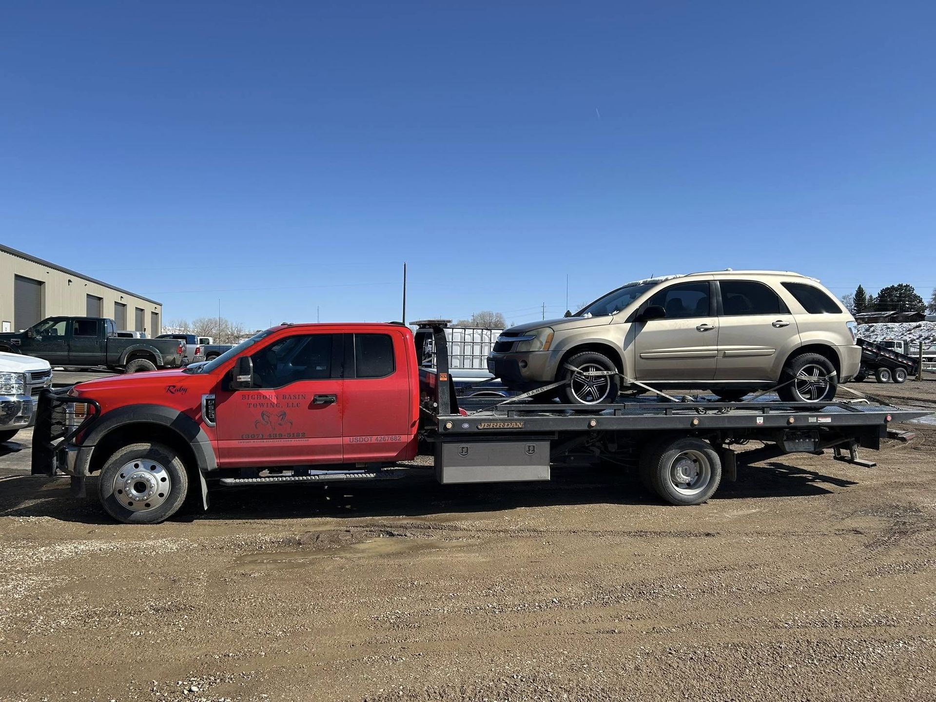 A red tow truck carries a gold SUV on its flatbed in a sunny outdoor setting.