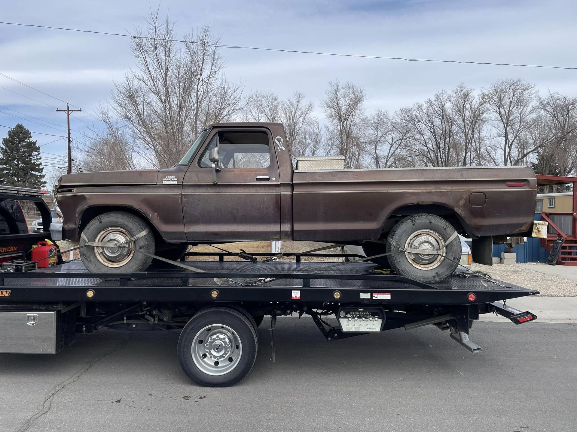 A brown, weathered pickup truck is secured on a black flatbed tow truck in a daytime setting.