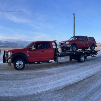 Red tow truck carrying a damaged red pickup truck on a snowy road with mountains in the background.