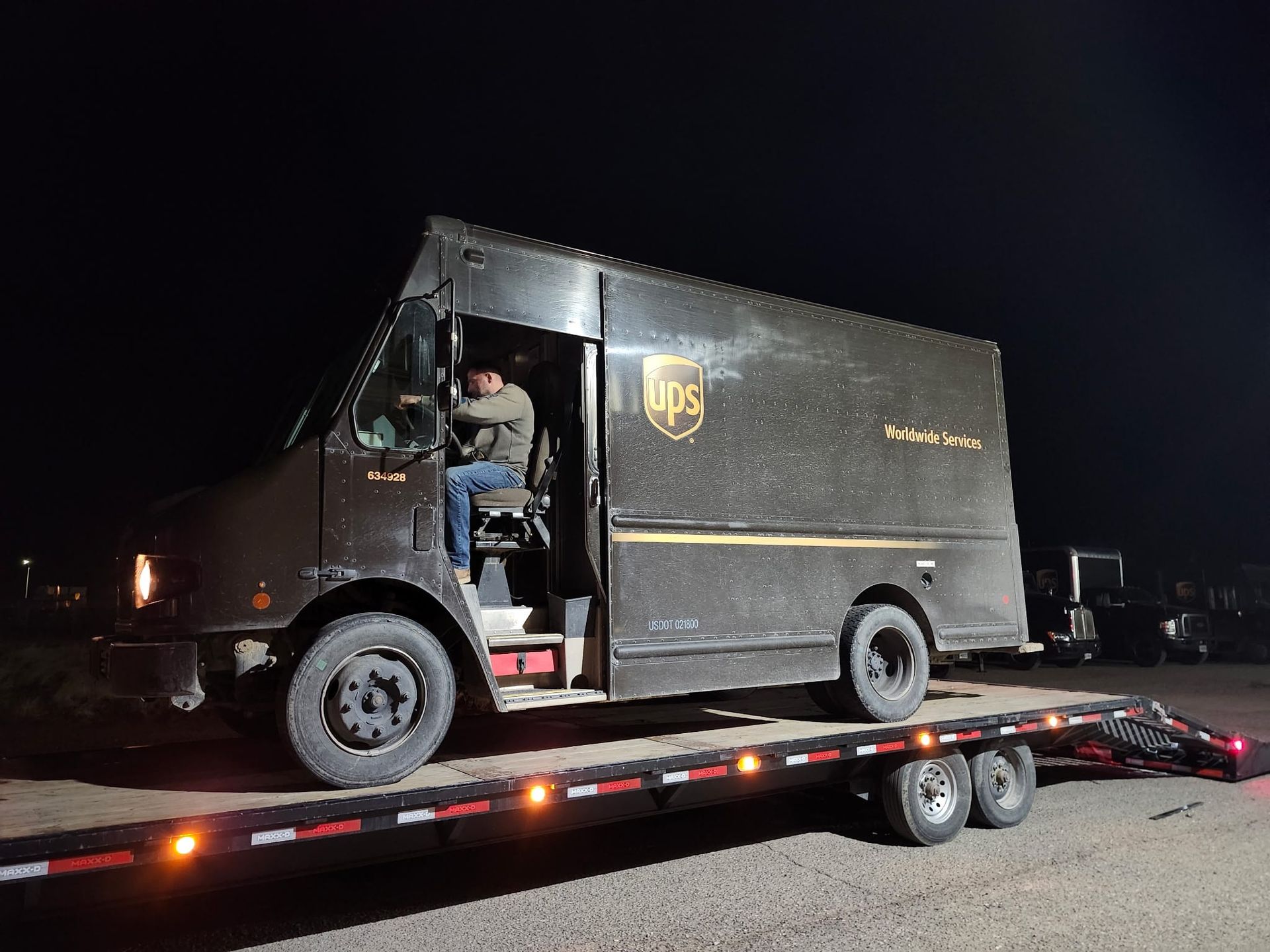 UPS truck on a flatbed trailer at night. Driver sits in the cab. Brown vehicle on a dark background.