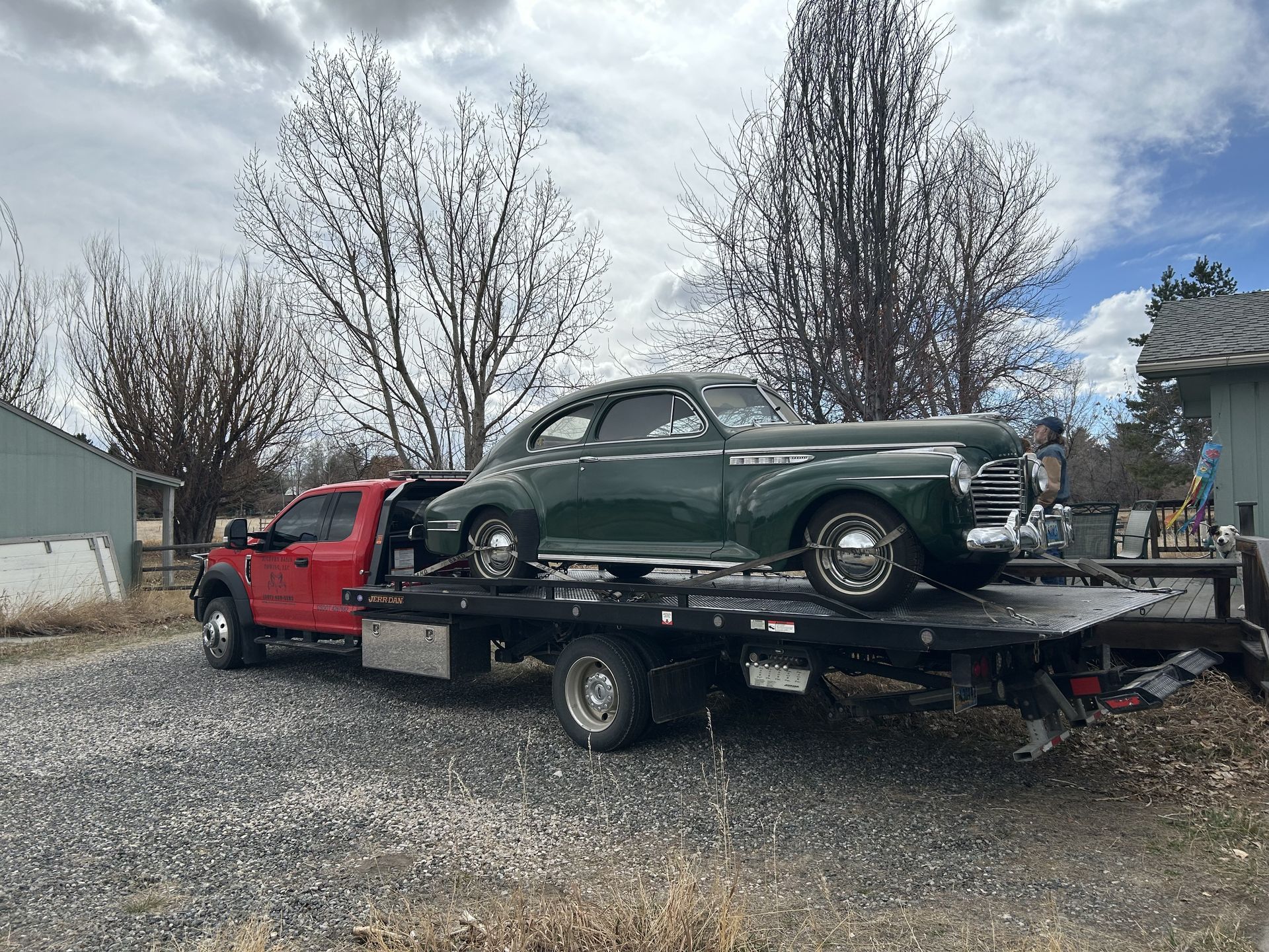 A vintage green car is loaded onto a red flatbed tow truck in a rural area. The sky is overcast.