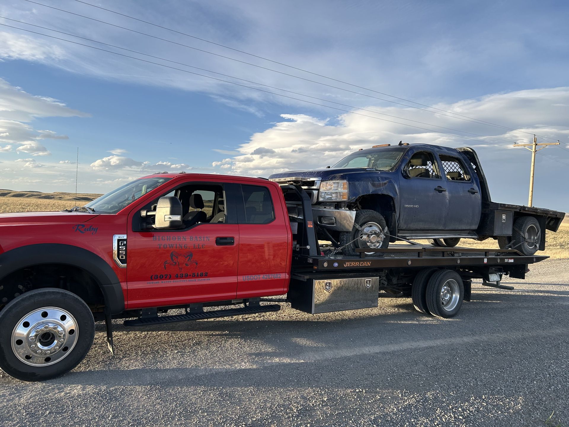 Red tow truck hauling a blue pickup truck on a flatbed trailer on a road under a partly cloudy sky.