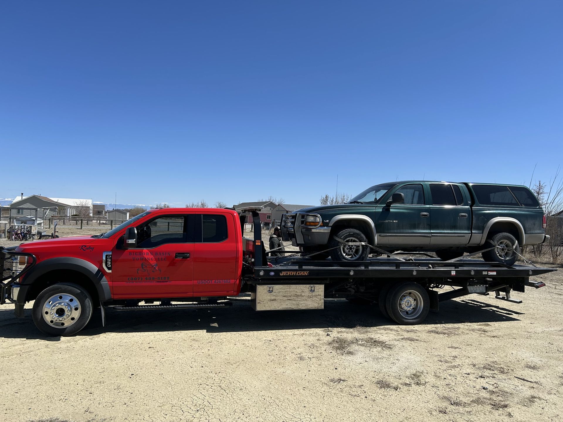 Red tow truck carrying a green pickup truck on its flatbed, outdoors under a blue sky.