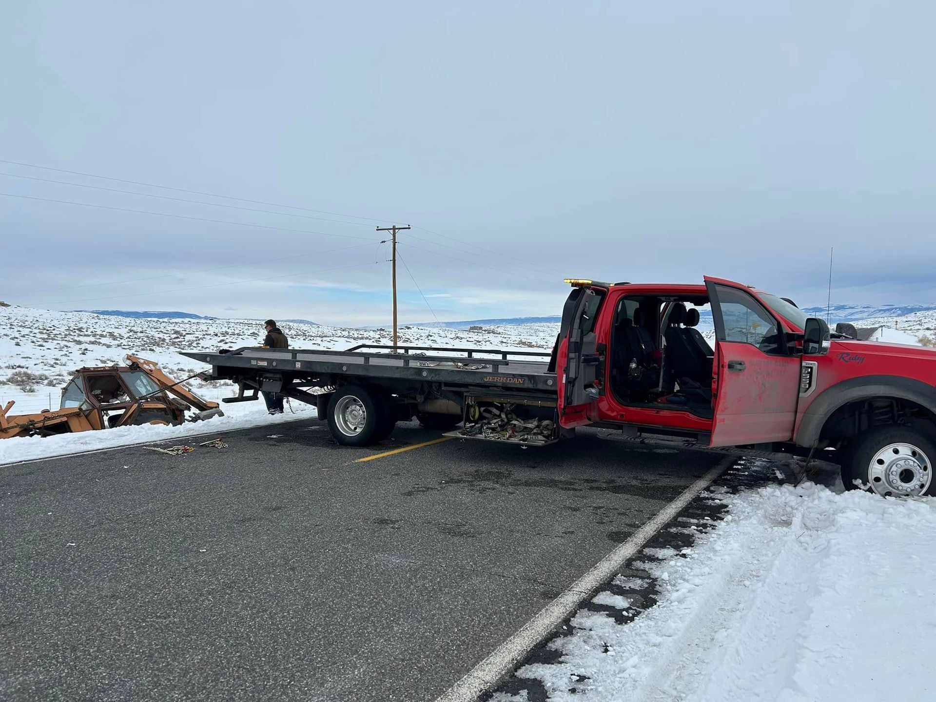 A red tow truck with its door open is stuck in snow beside a highway. A damaged vehicle sits on its bed.