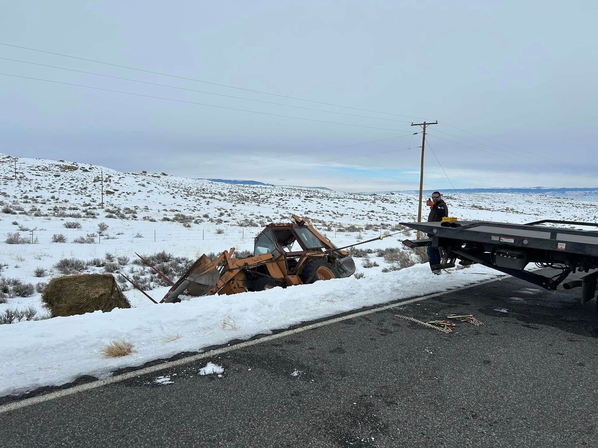 An orange tractor is stuck in snow beside a road. A tow truck prepares to pull it out. Snowy landscape.