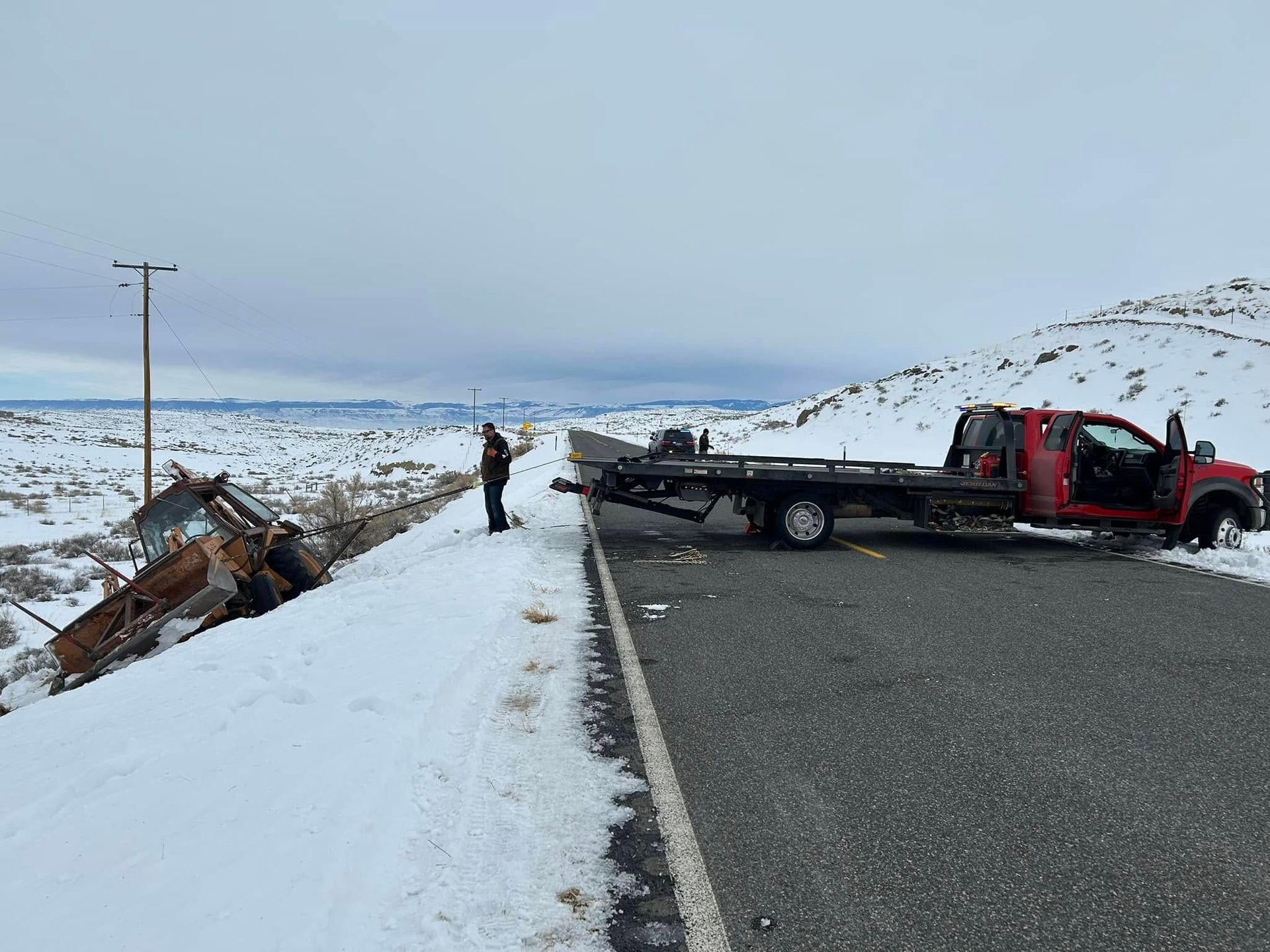 A tow truck at the side of a snow-covered road near a damaged vehicle on a hillside. A person stands next to the truck.