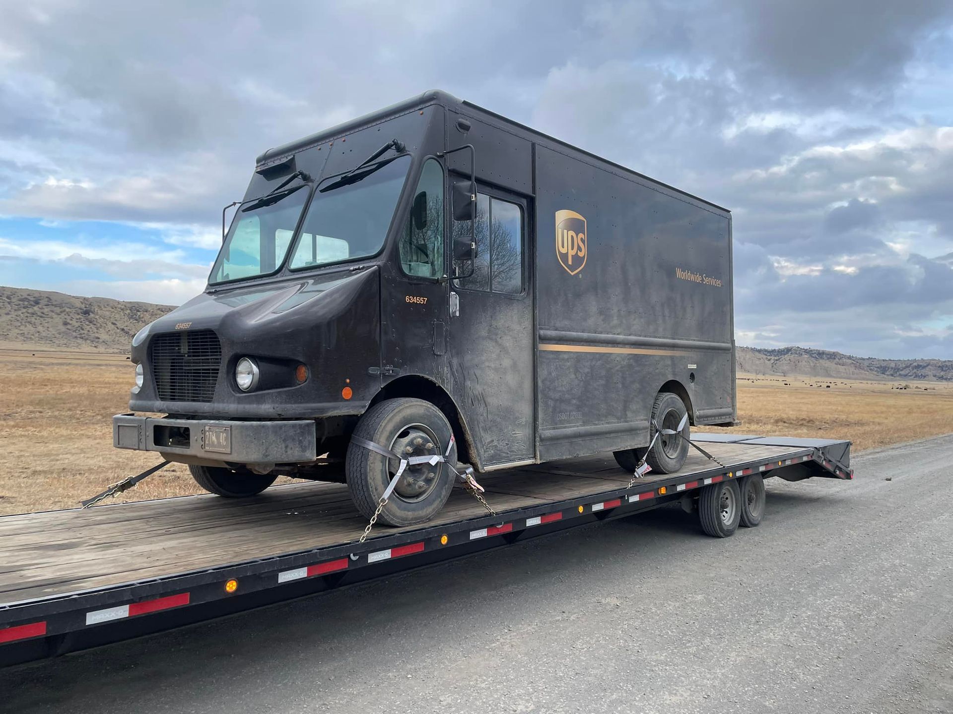A UPS truck loaded onto a flatbed trailer on a rural road. The truck is dark brown and has the UPS logo. The sky is overcast.