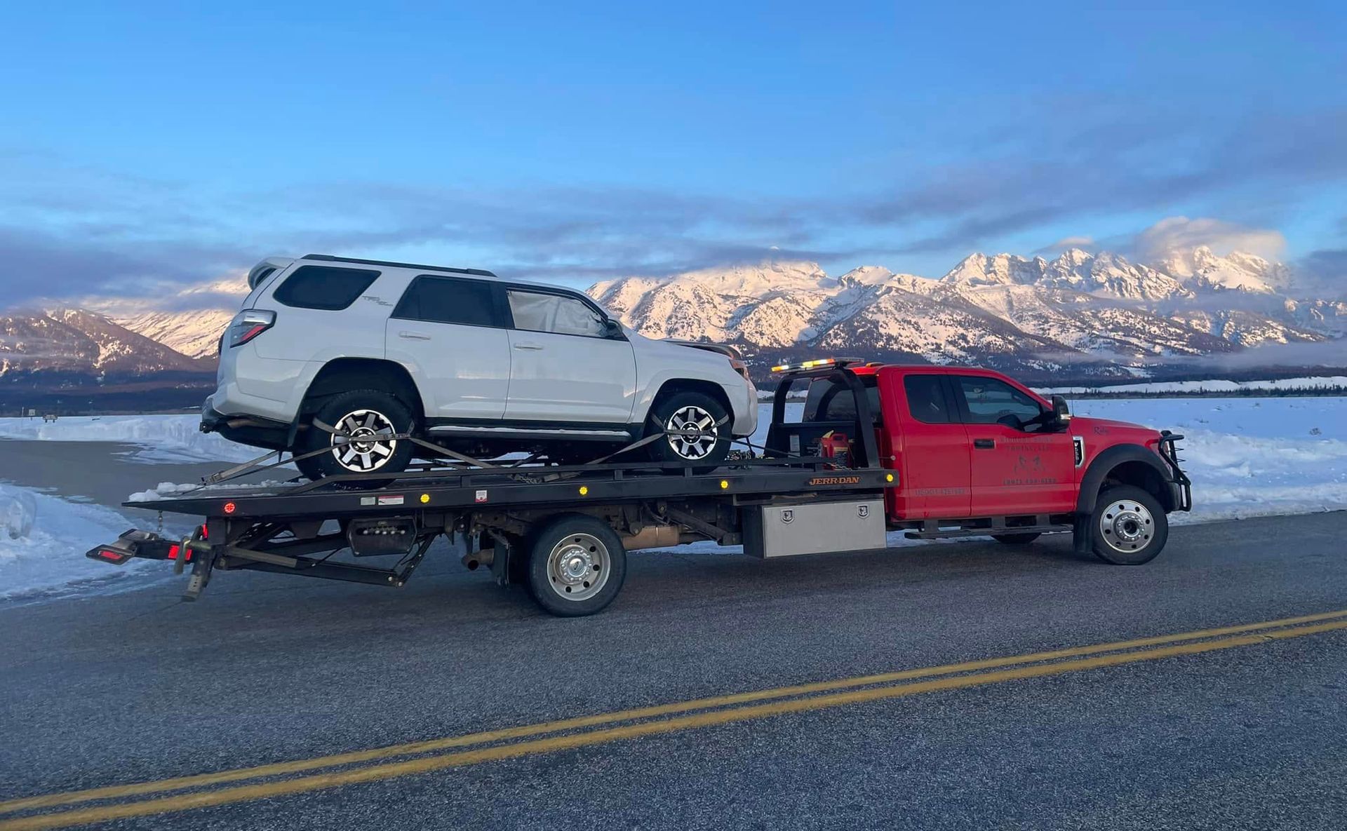 A white SUV loaded onto a red tow truck on a road, mountains in the background. Snowy scene.