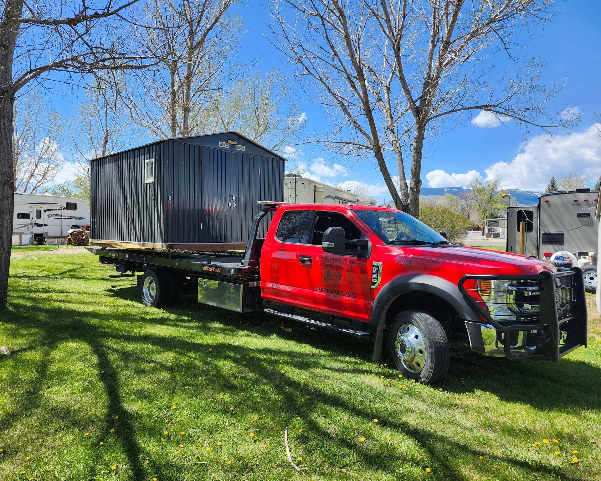 Red flatbed truck carrying a dark storage container on a grassy field near trees and RVs.