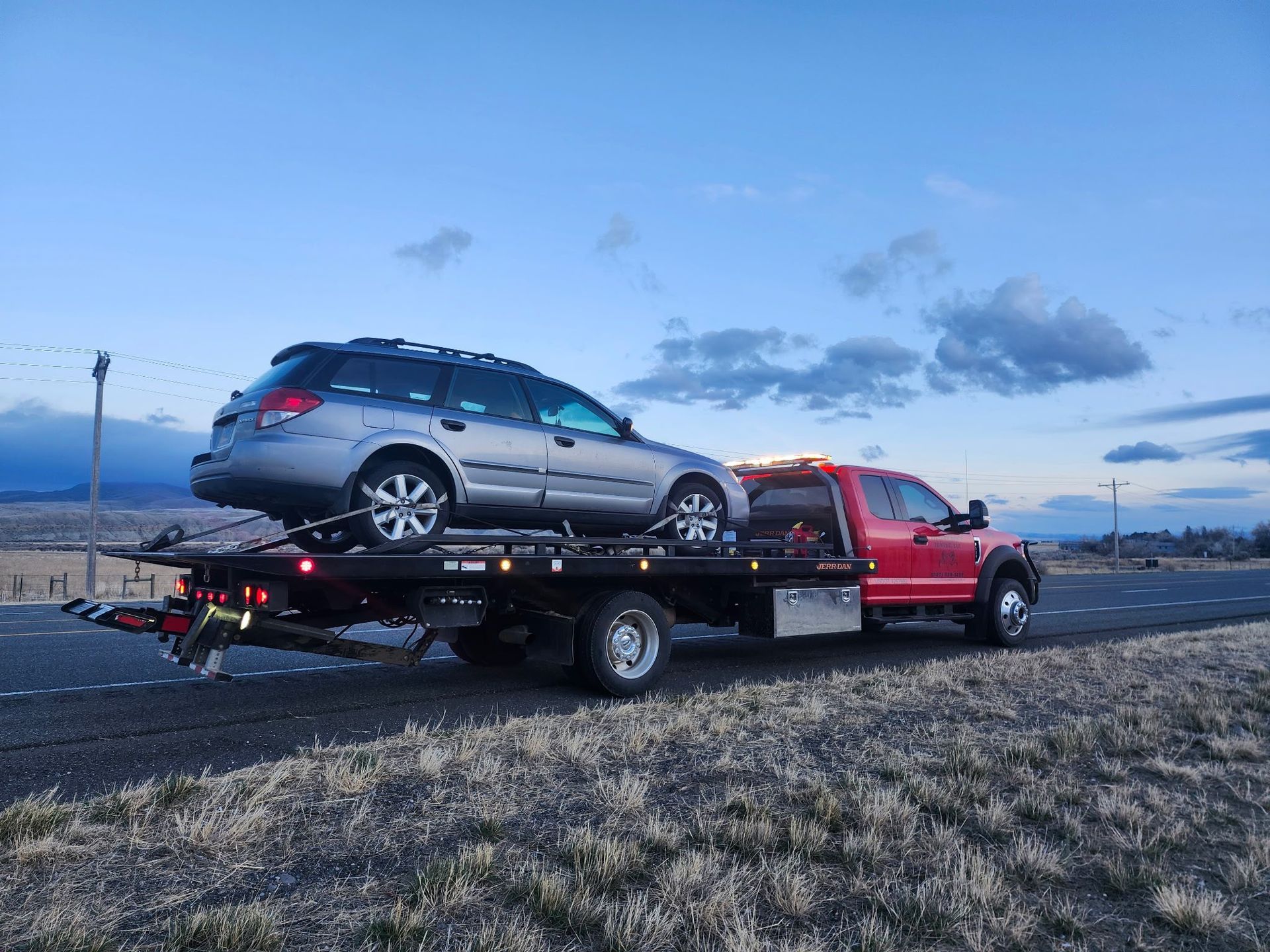 A red tow truck carrying a silver station wagon on its flatbed on a road at dusk.