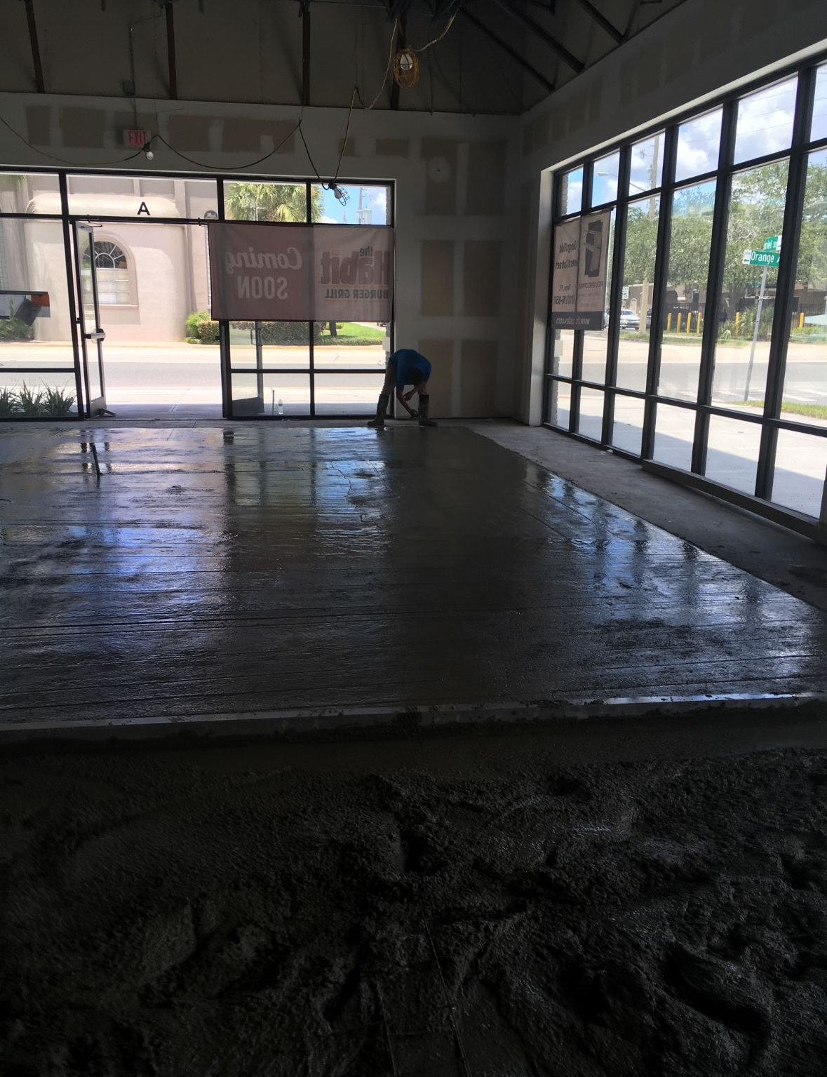 Construction worker smoothing wet cement floor in a building with large windows.