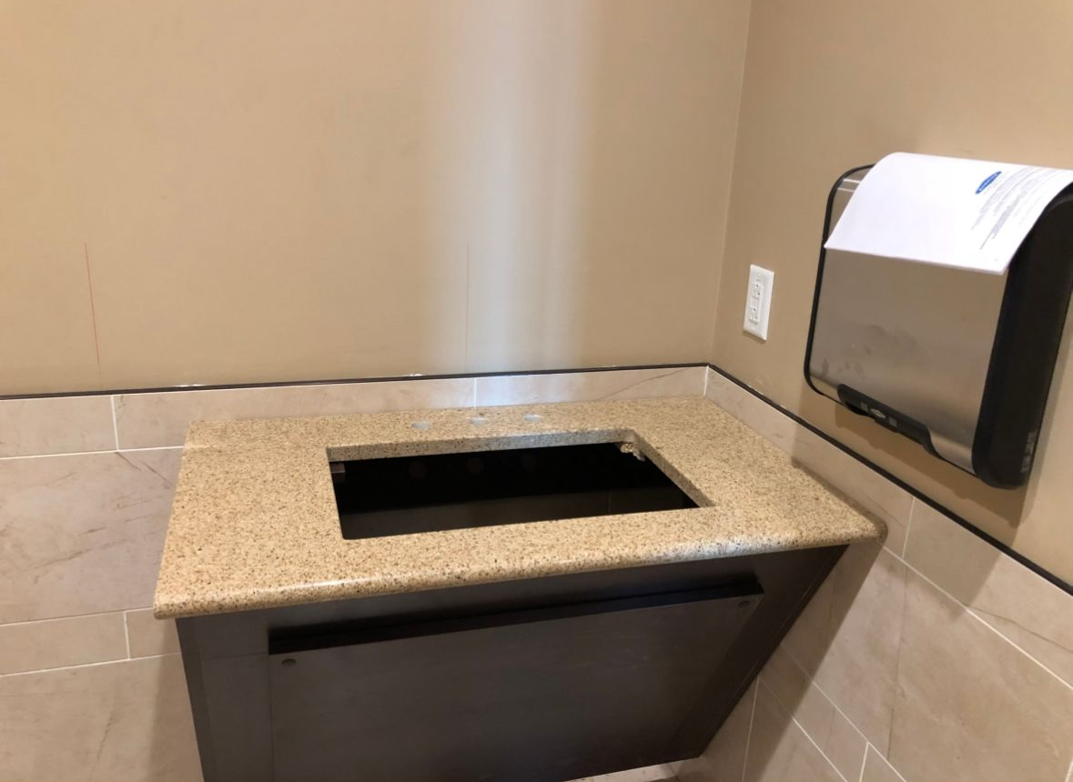Public restroom with an empty sink and paper towel dispenser. Beige walls and countertops.