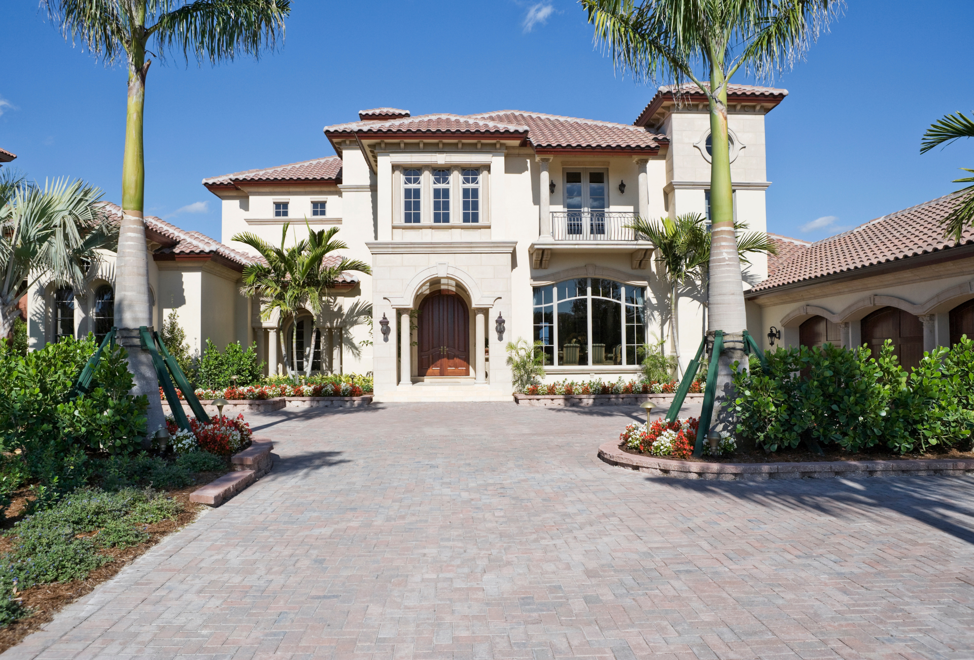 Elegant beige mansion with tile roof, palm trees, and brick driveway on a sunny day.