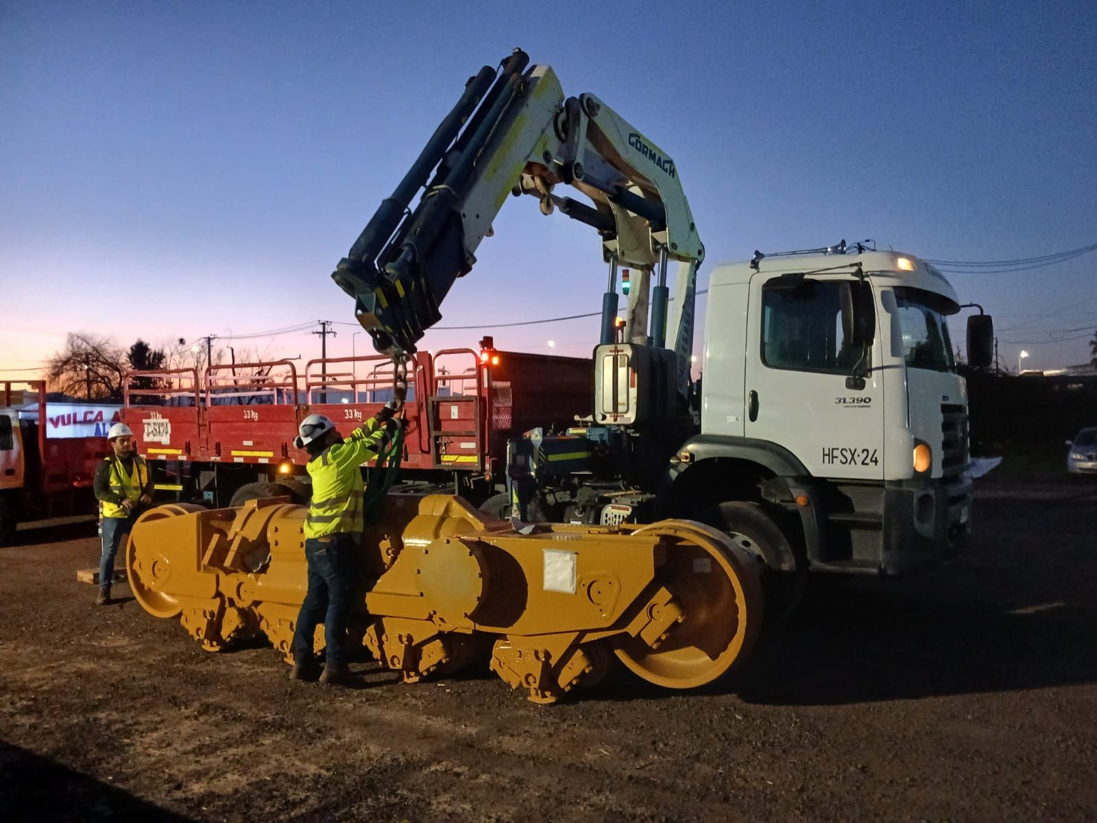 Trabajadores utilizan una grúa para cargar equipos de construcción de carreteras en un camión al anochecer.