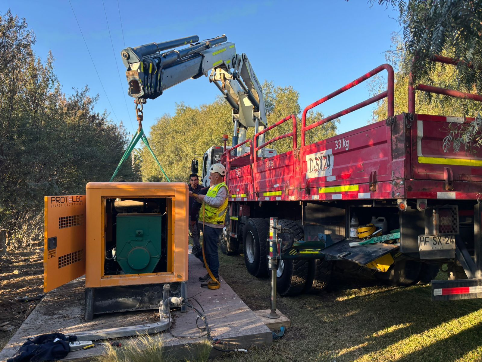 Una grúa levanta un generador amarillo de la plataforma de un camión; dos trabajadores permanecen cerca, afuera.