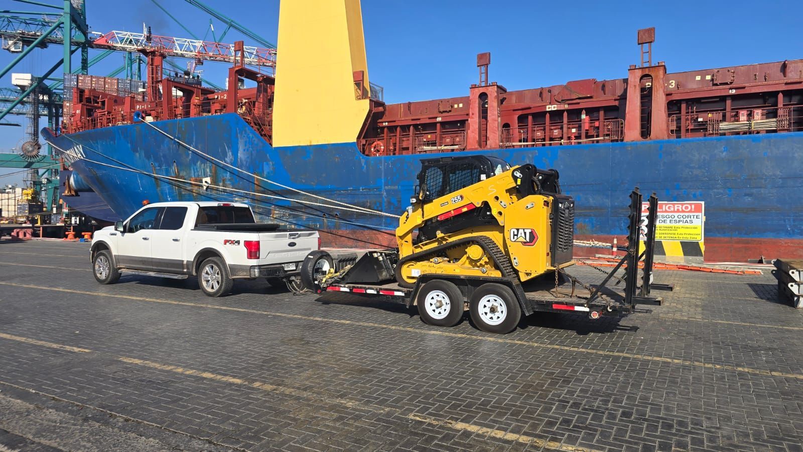 Camión blanco remolcando una minicargadora amarilla sobre un remolque en un puerto, con un gran barco en el fondo.