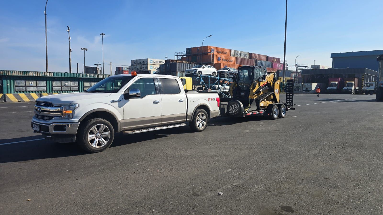 Camioneta blanca remolcando equipo en un puerto, con contenedores de envío al fondo.