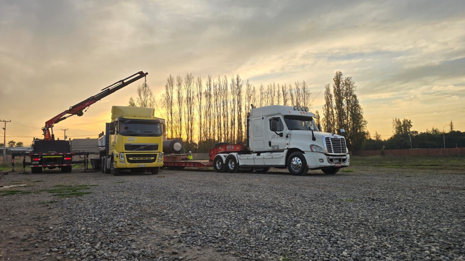 Camiones estacionados sobre grava al atardecer, uno de ellos con grúa.