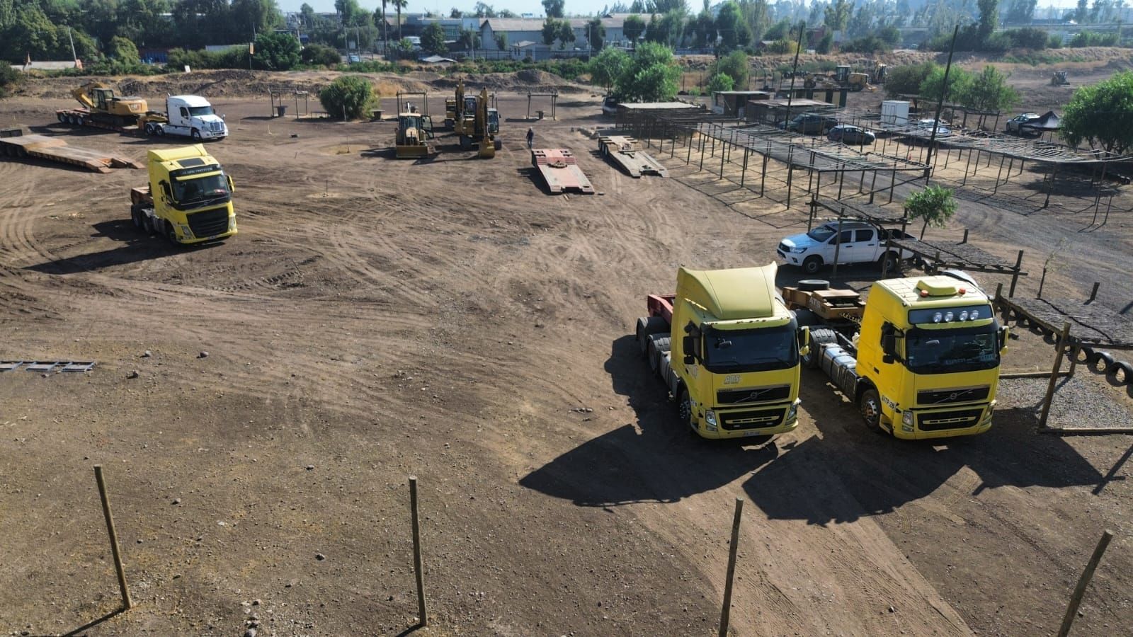Tres camiones semirremolque amarillos estacionados en un lote de tierra con equipos de construcción y una camioneta blanca al fondo.