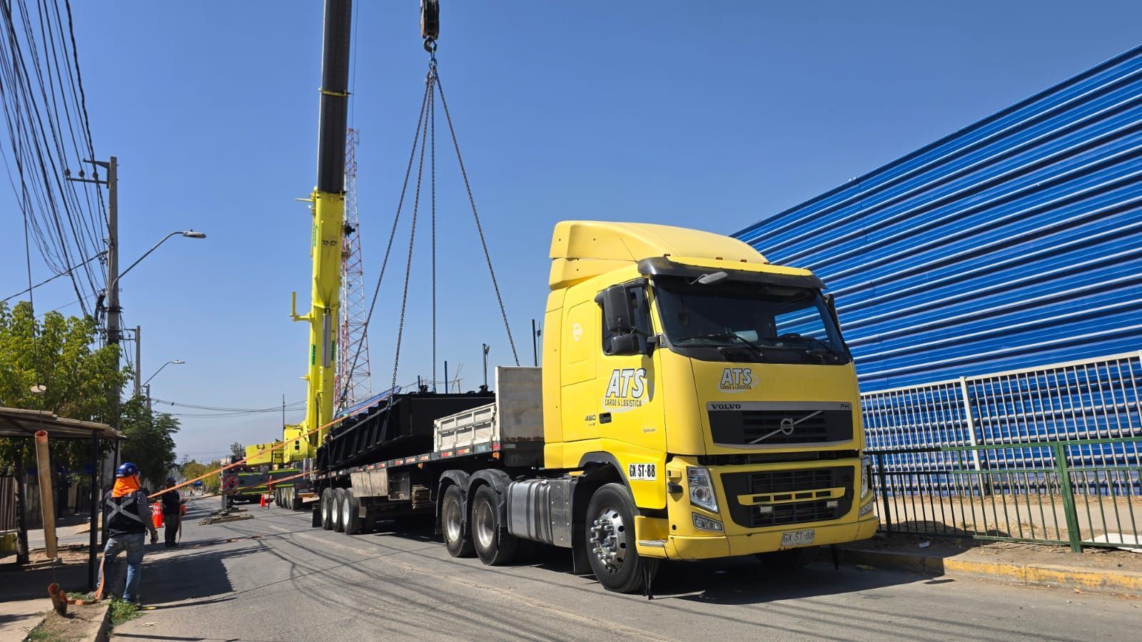 Camión semirremolque amarillo con grúa cargando materiales en una calle soleada; trabajadores de la construcción presentes.