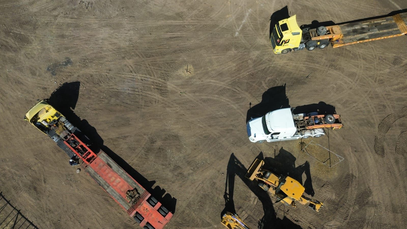 Vista aérea de vehículos de construcción: camiones amarillos y blancos con remolques, excavadora amarilla sobre tierra.