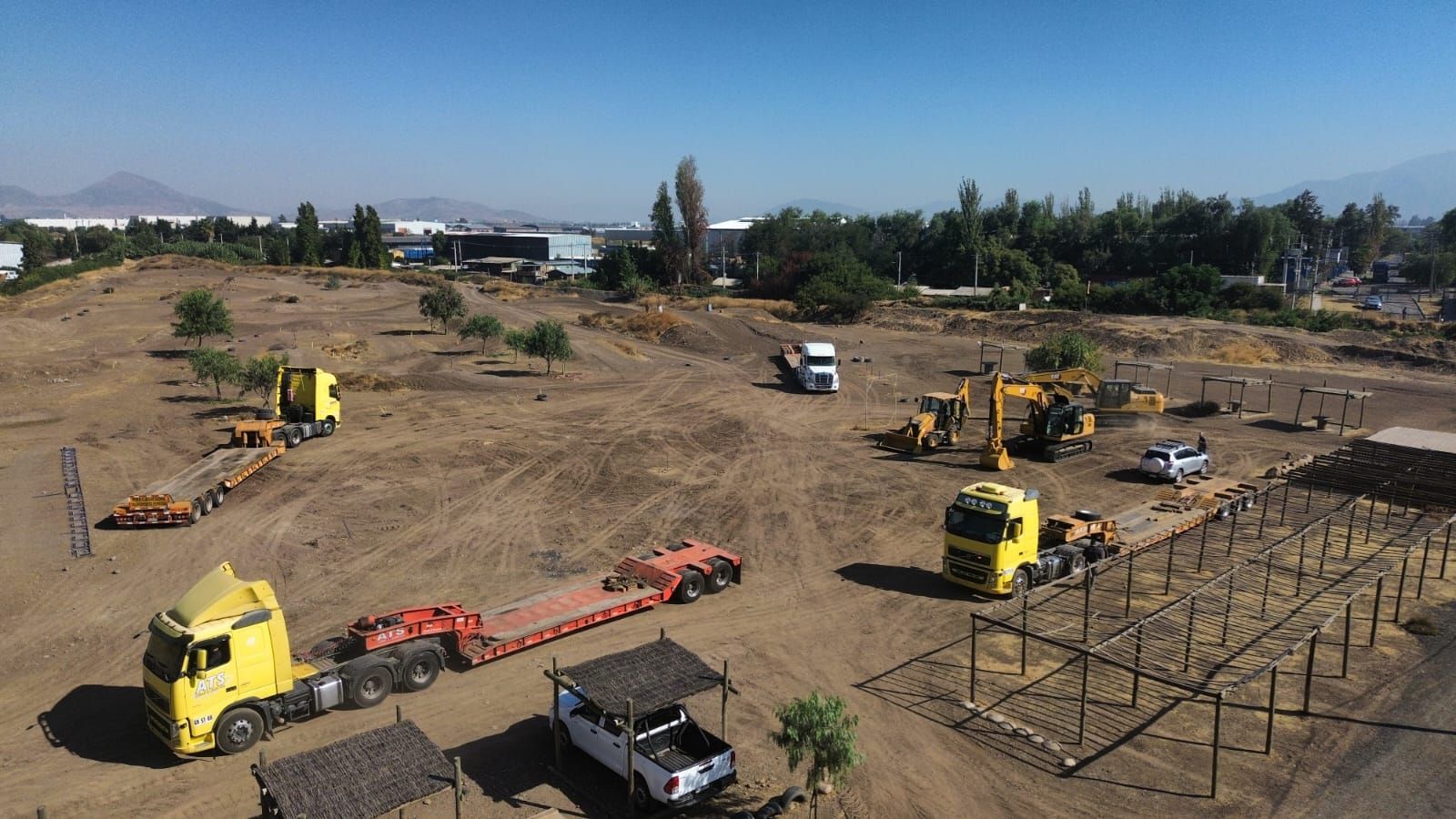 Sitio de construcción con camiones amarillos y maquinaria pesada en un lote de tierra, cielo azul.