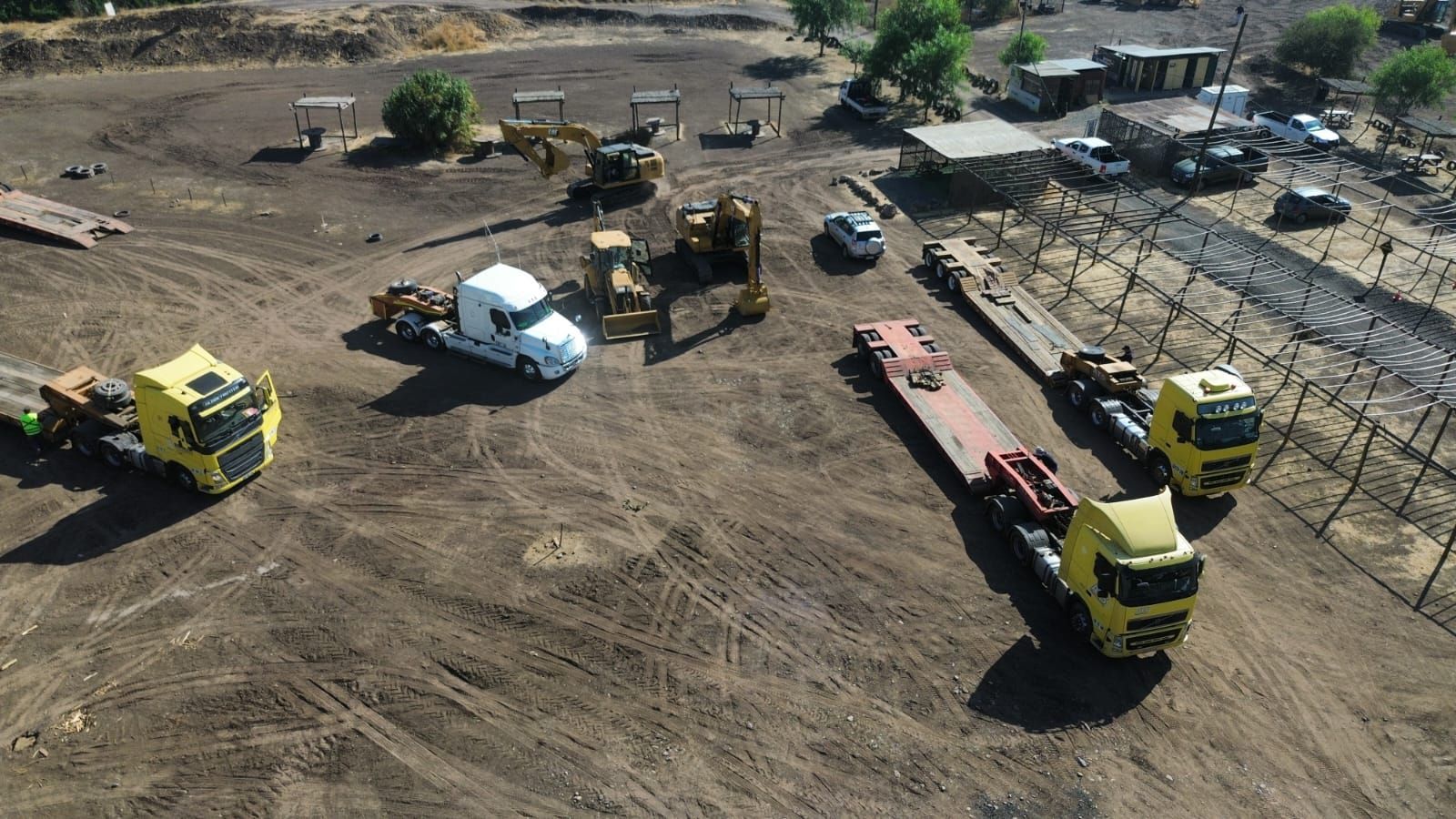 Vista aérea: Sitio de construcción con camiones, maquinaria pesada y edificios; suelo de tierra marrón.