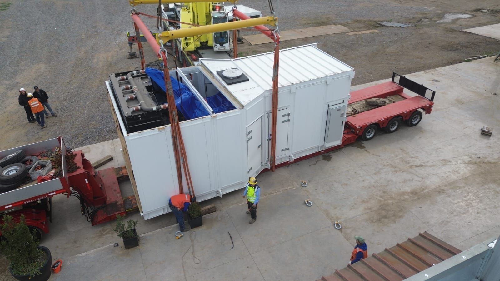 Un gran contenedor blanco es elevado con una grúa a un remolque. Trabajadores con chalecos de seguridad observan.