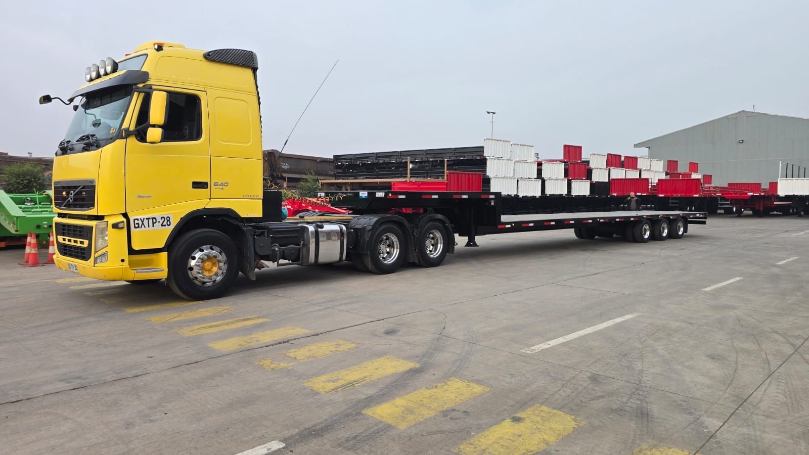 Camión semirremolque amarillo con plataforma cargada con pilas de objetos rojos y blancos en una zona pavimentada.