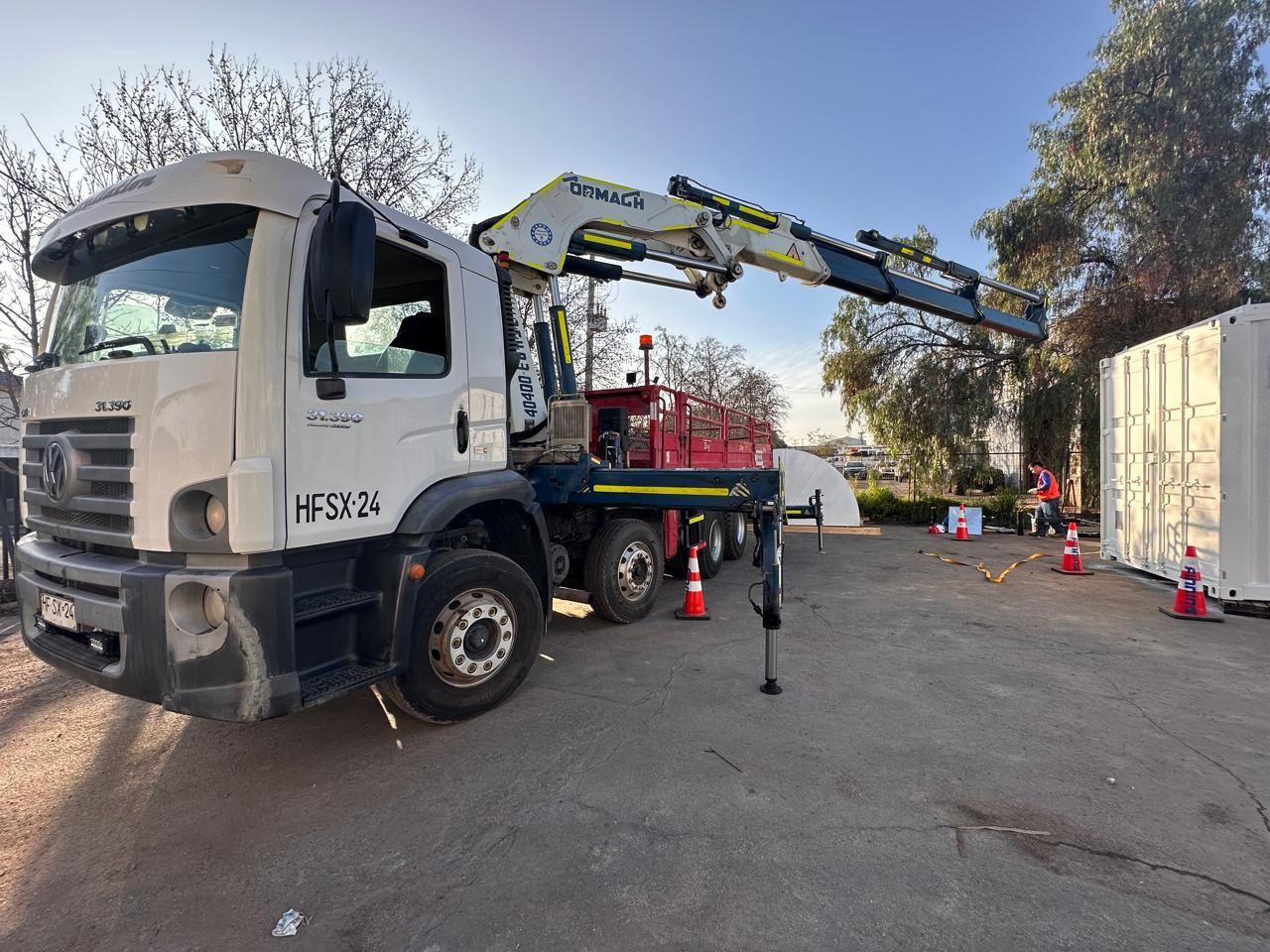 Camión blanco con grúa extendida, estacionado cerca de un contenedor blanco. Conos naranjas instalados.