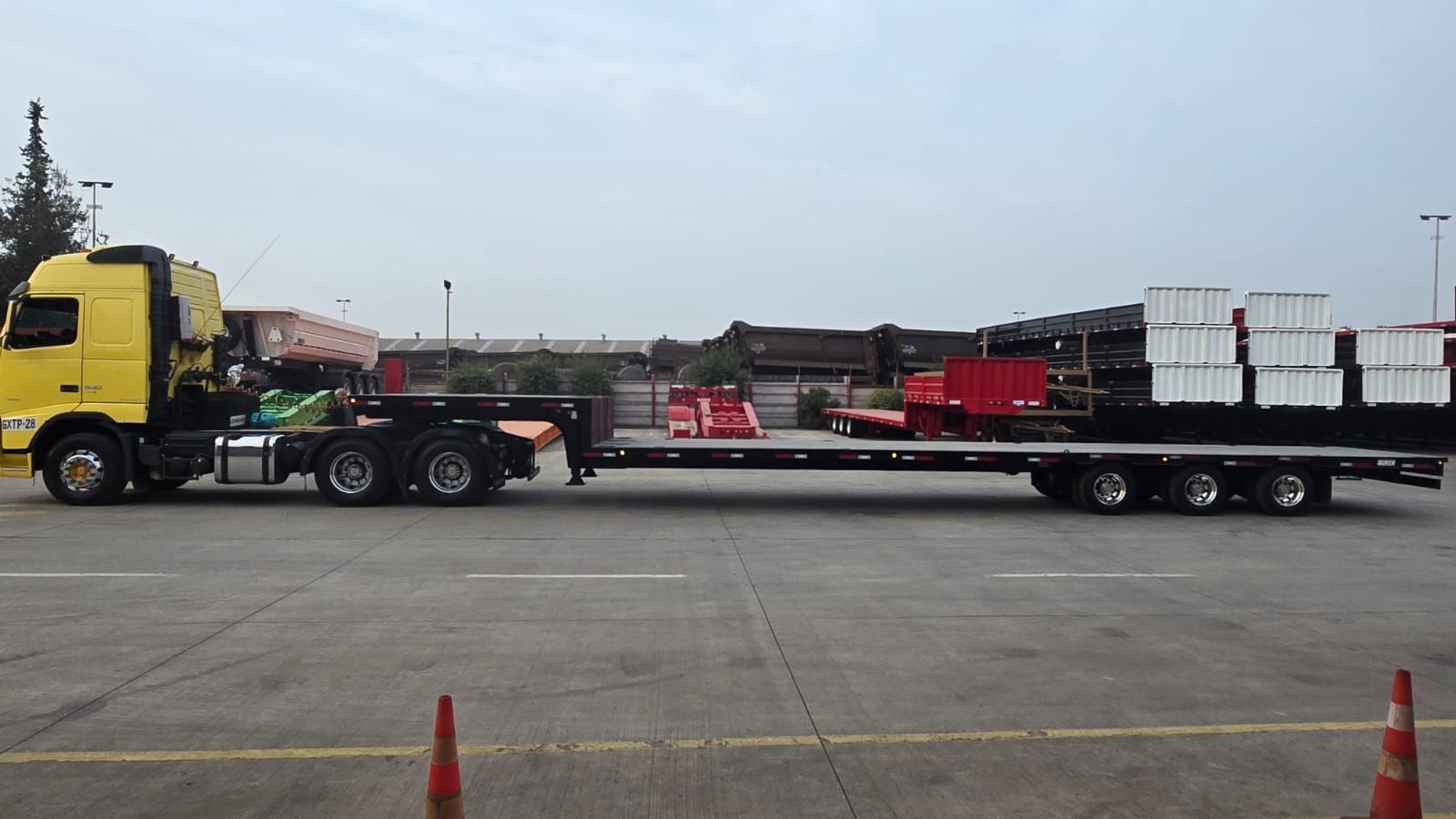 Camión semirremolque amarillo que transporta un remolque de plataforma cargado con cajas blancas y contenedores rojos sobre asfalto.