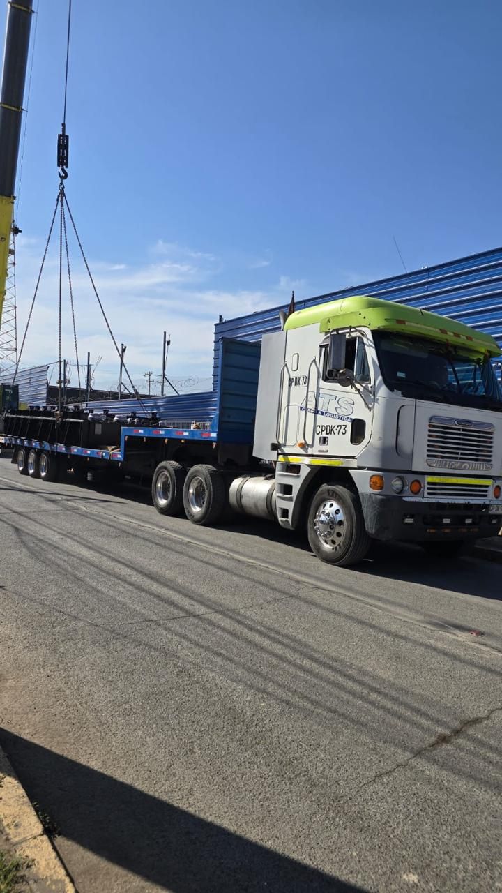 Un gran camión que transporta una plataforma pesada con carga es levantado por una grúa en un día soleado.