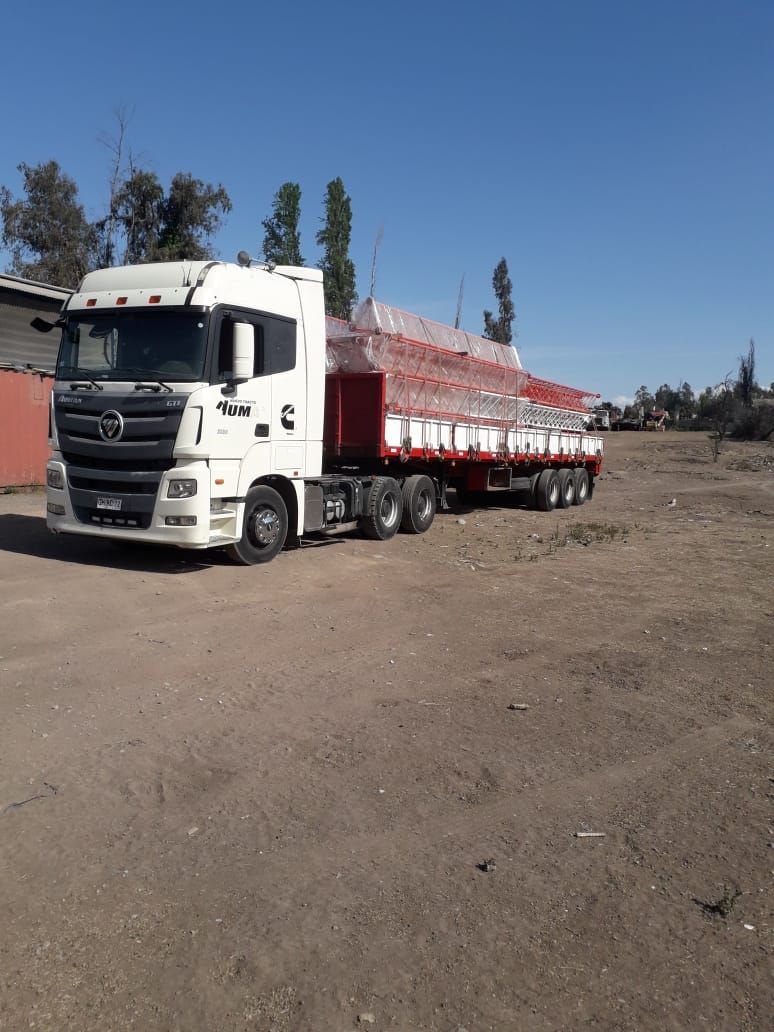 Camión semirremolque blanco con remolque rojo que transporta carga en un terreno de tierra bajo un cielo azul.