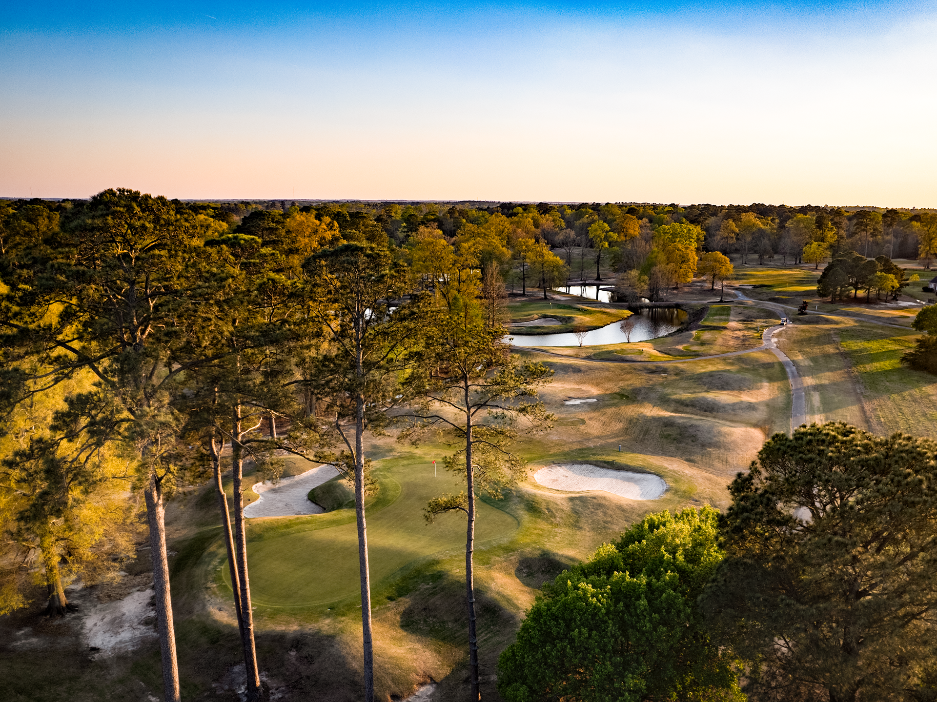 An aerial view of a golf course surrounded by trees at sunset. An aerial view of a golf course surrounded by trees at sunset.