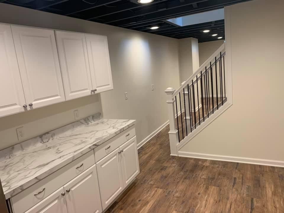 White cabinets with a marble countertop in a basement, beside a staircase with black spindles.