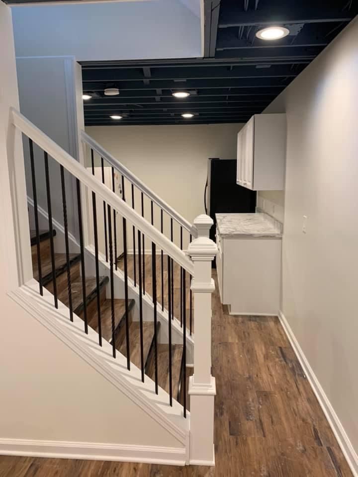 Staircase leading to a kitchenette in a finished basement. White walls and cabinets, black ceiling, wood-look floors.