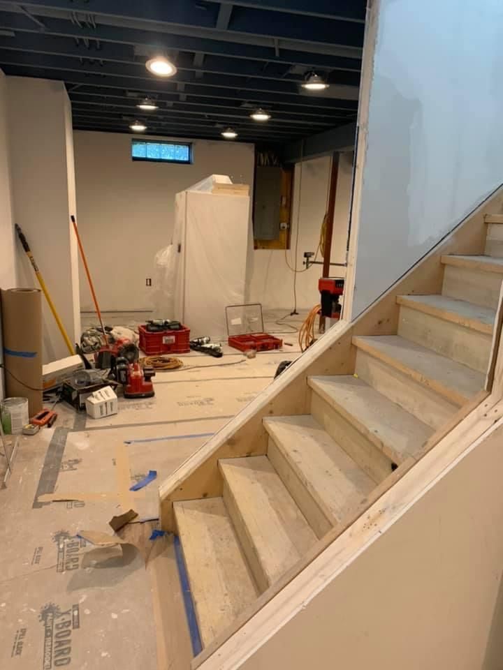 Unfinished basement with stairs, tools, and construction materials. Beige walls, dark ceiling, and natural wood stairs.