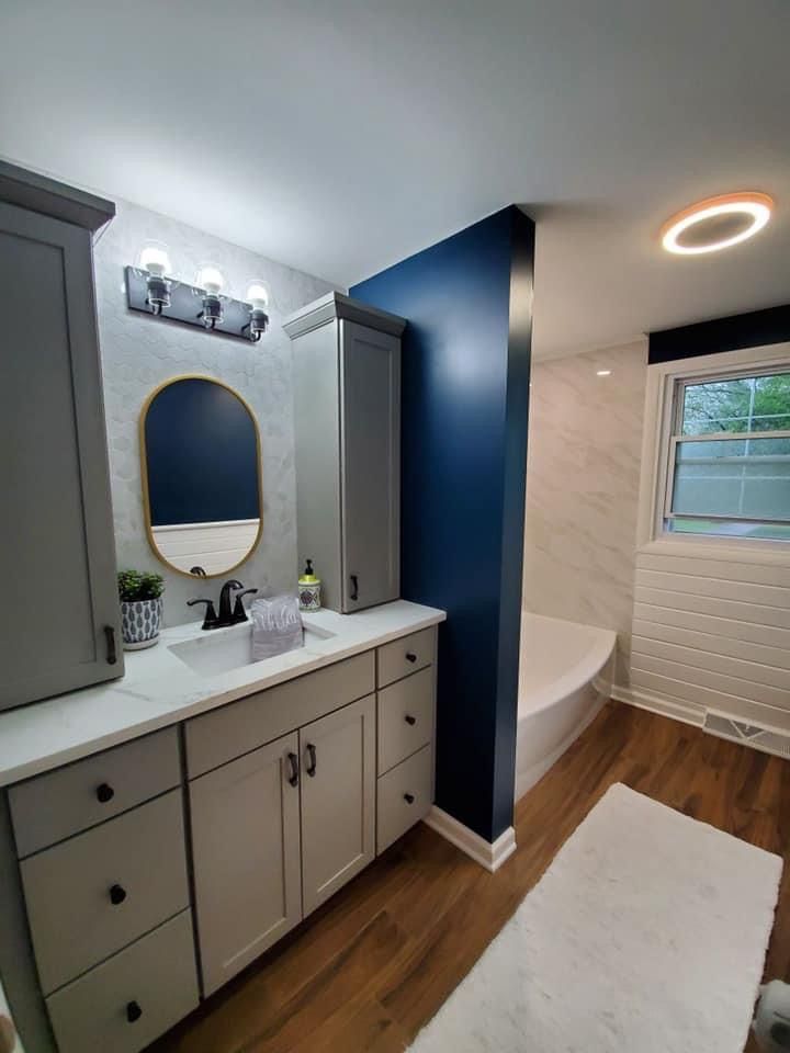 Gray and blue modern bathroom with cabinetry, mirror, tub, and hardwood floor.