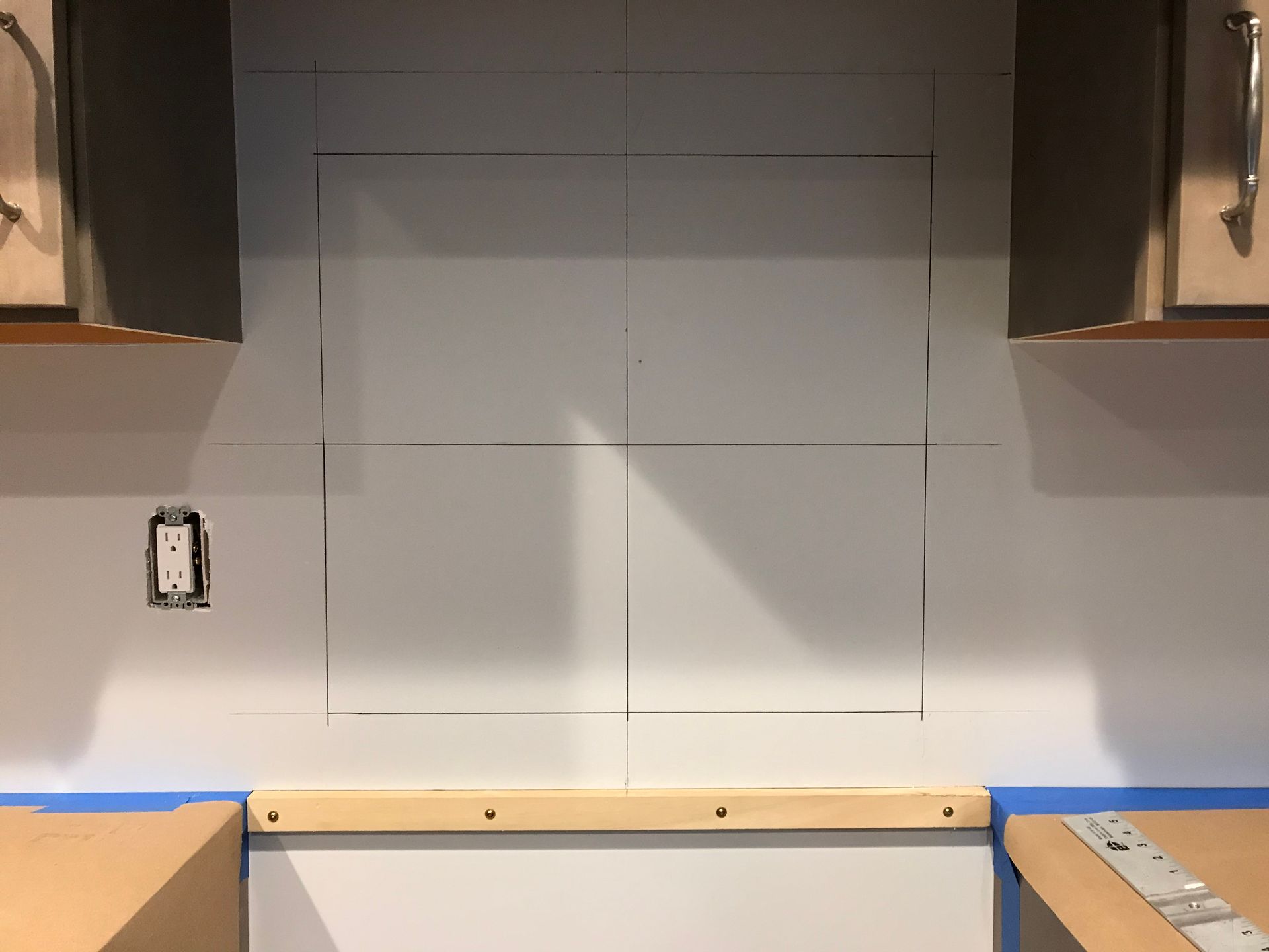 A kitchen wall with taped off areas for backsplash, with cabinet edges and electrical outlet visible.