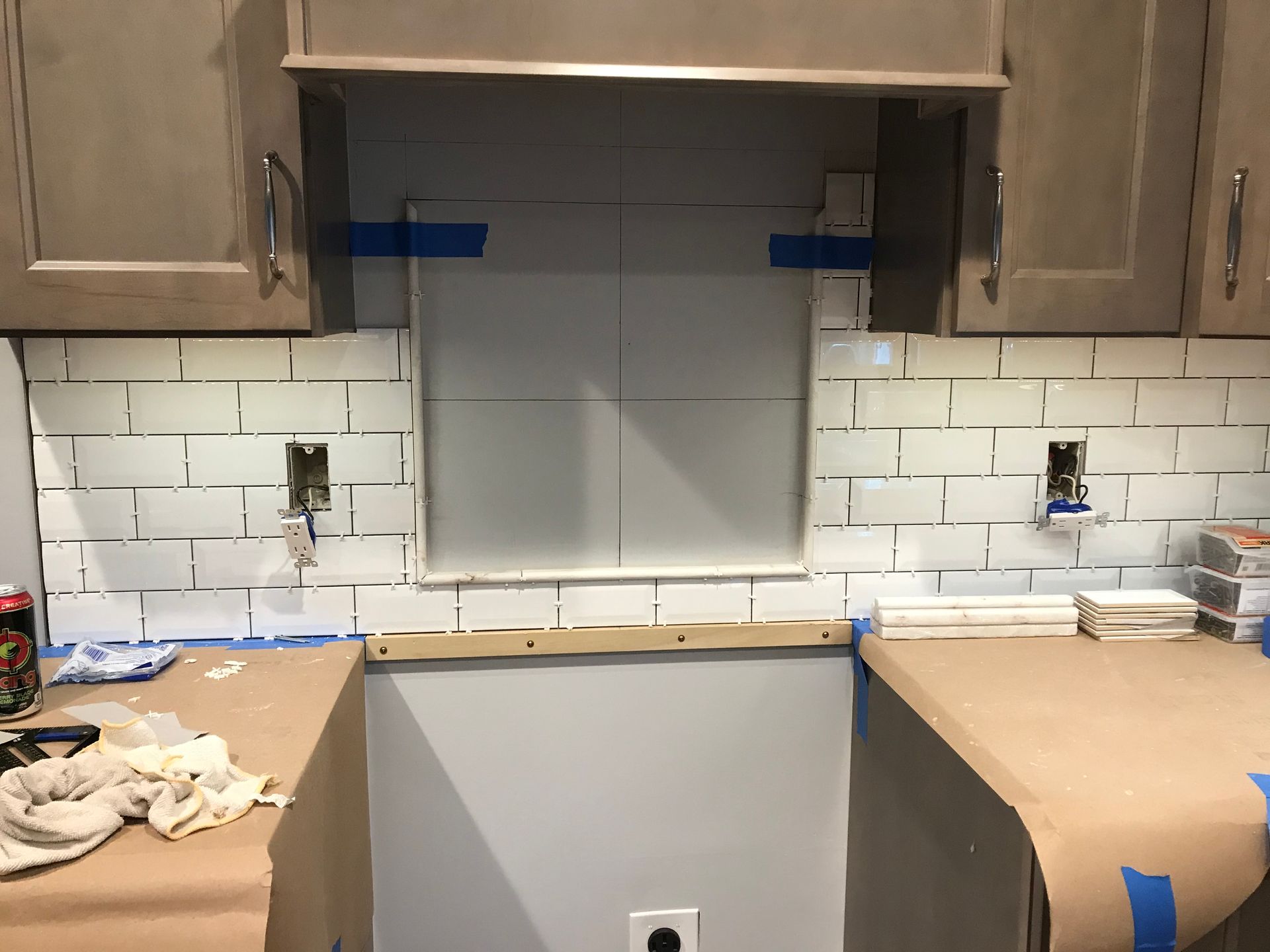 Kitchen backsplash installation in progress; white subway tiles, gray cabinets, countertops covered in brown paper.