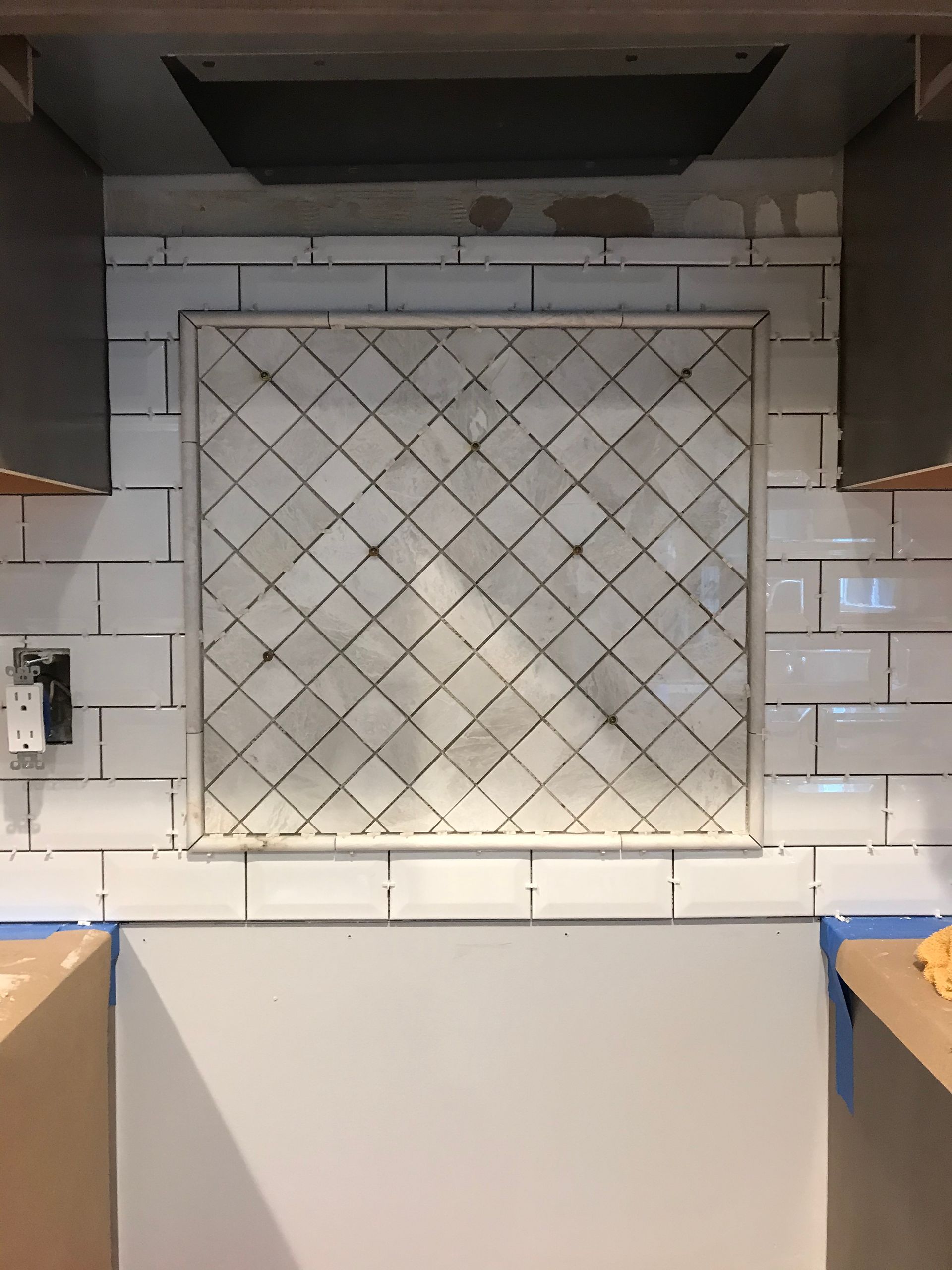 Kitchen backsplash under construction: white subway tiles and diamond-patterned tile focal point, cabinets on sides.
