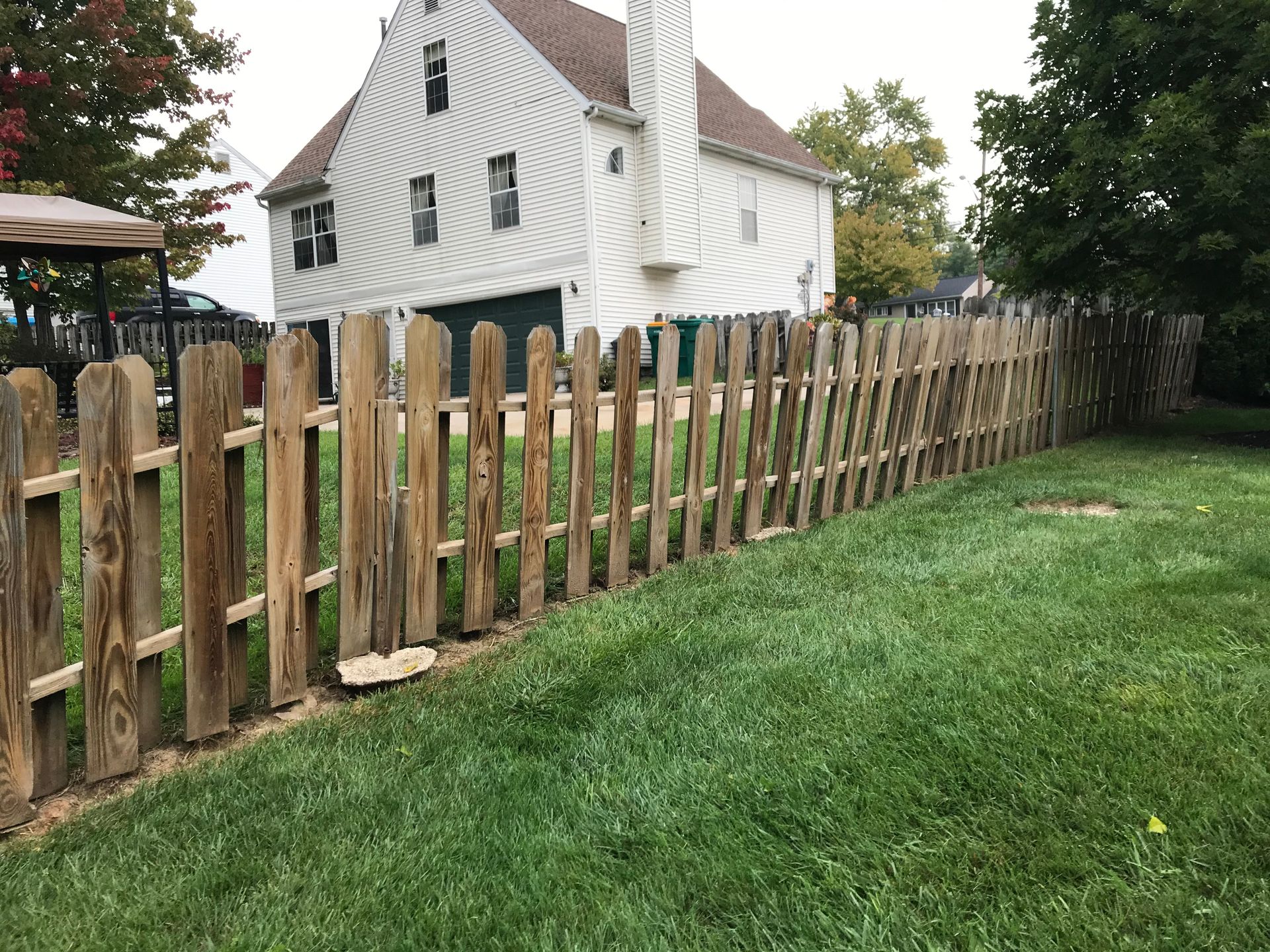 Wooden picket fence along green grass in front of a white house with a brown roof.
