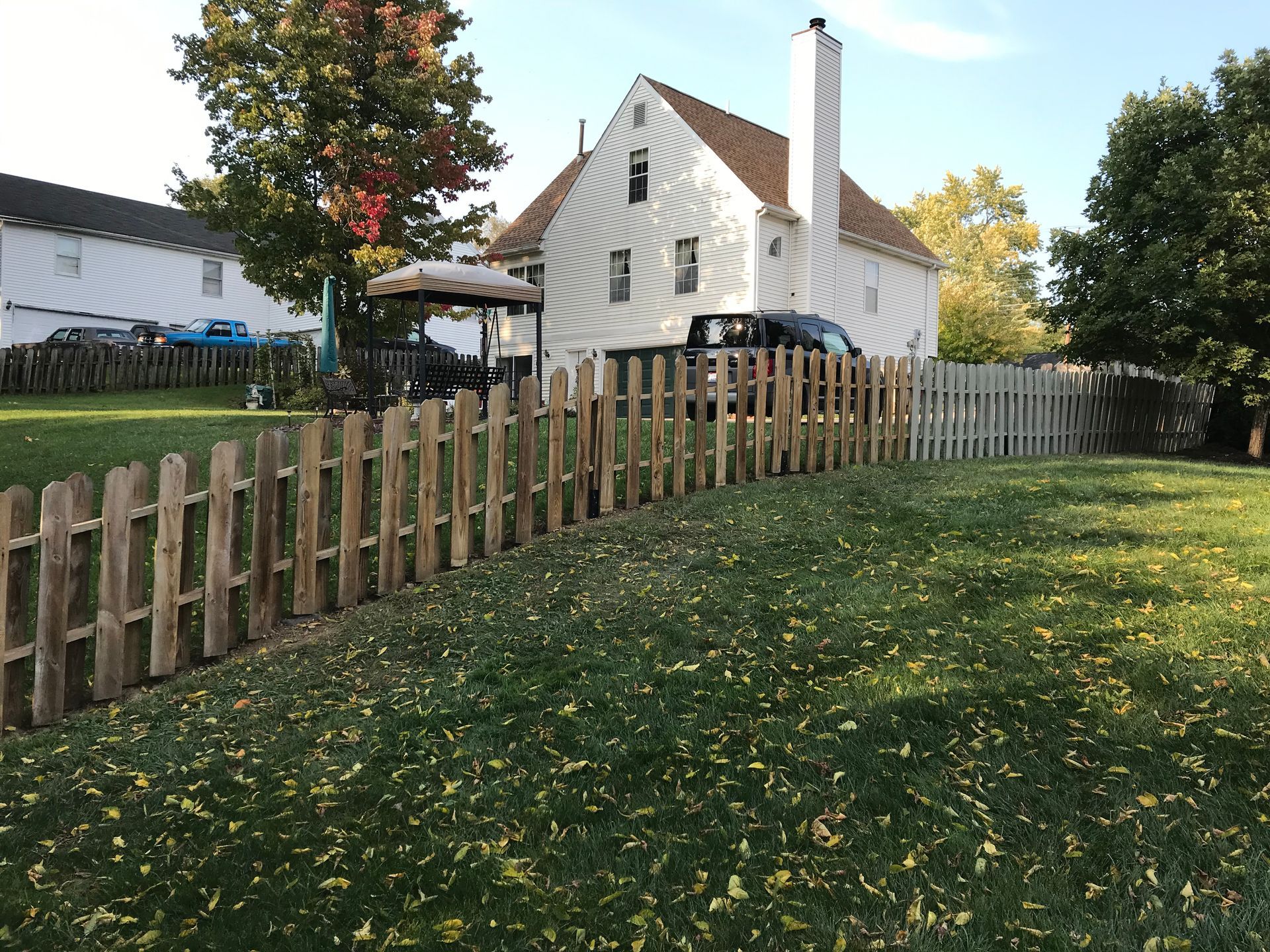 Wooden picket fence in front yard, a two-story house and trees in the background on a sunny day.