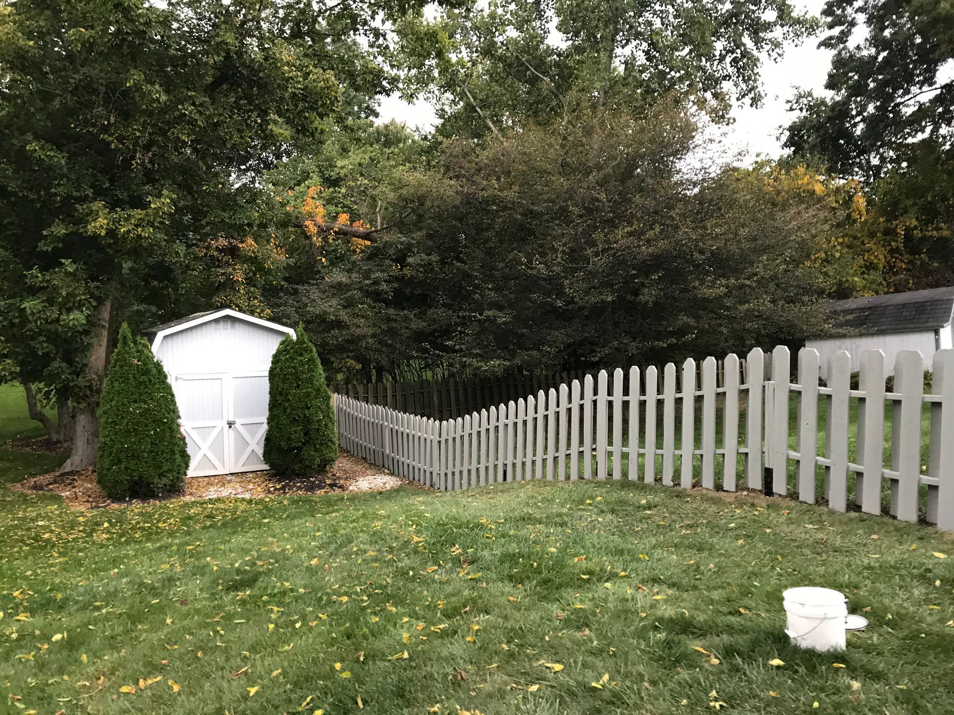 White shed and picket fence in backyard, green grass, trees in background.